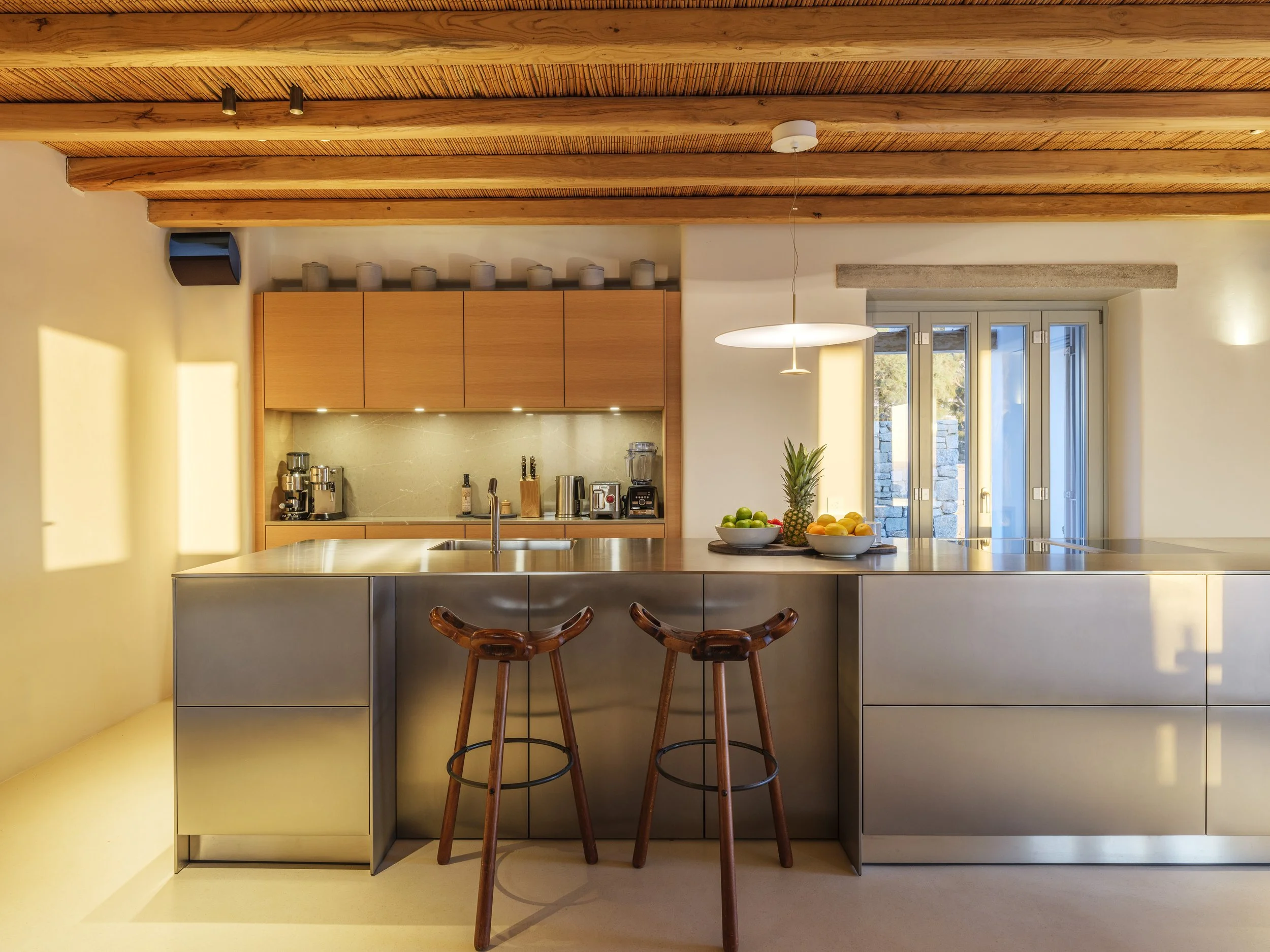 Modern kitchen with wood beams on the ceiling, a kitchen island with a light-colored surface, small bowls of fruits including pineapples and oranges, wooden bar stools, and a large window that lets in natural light.
