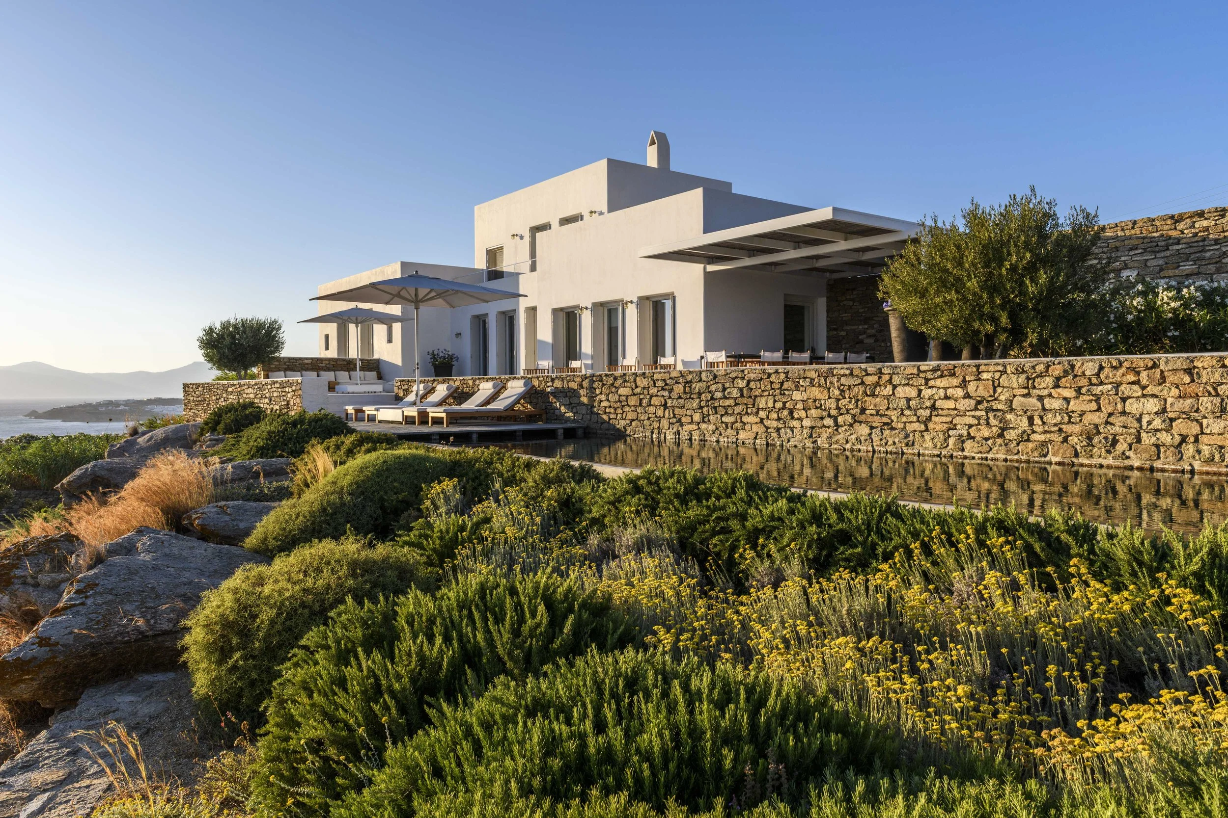 Modern white house with outdoor seating, umbrellas, and greenery situated near a body of water under a clear blue sky.