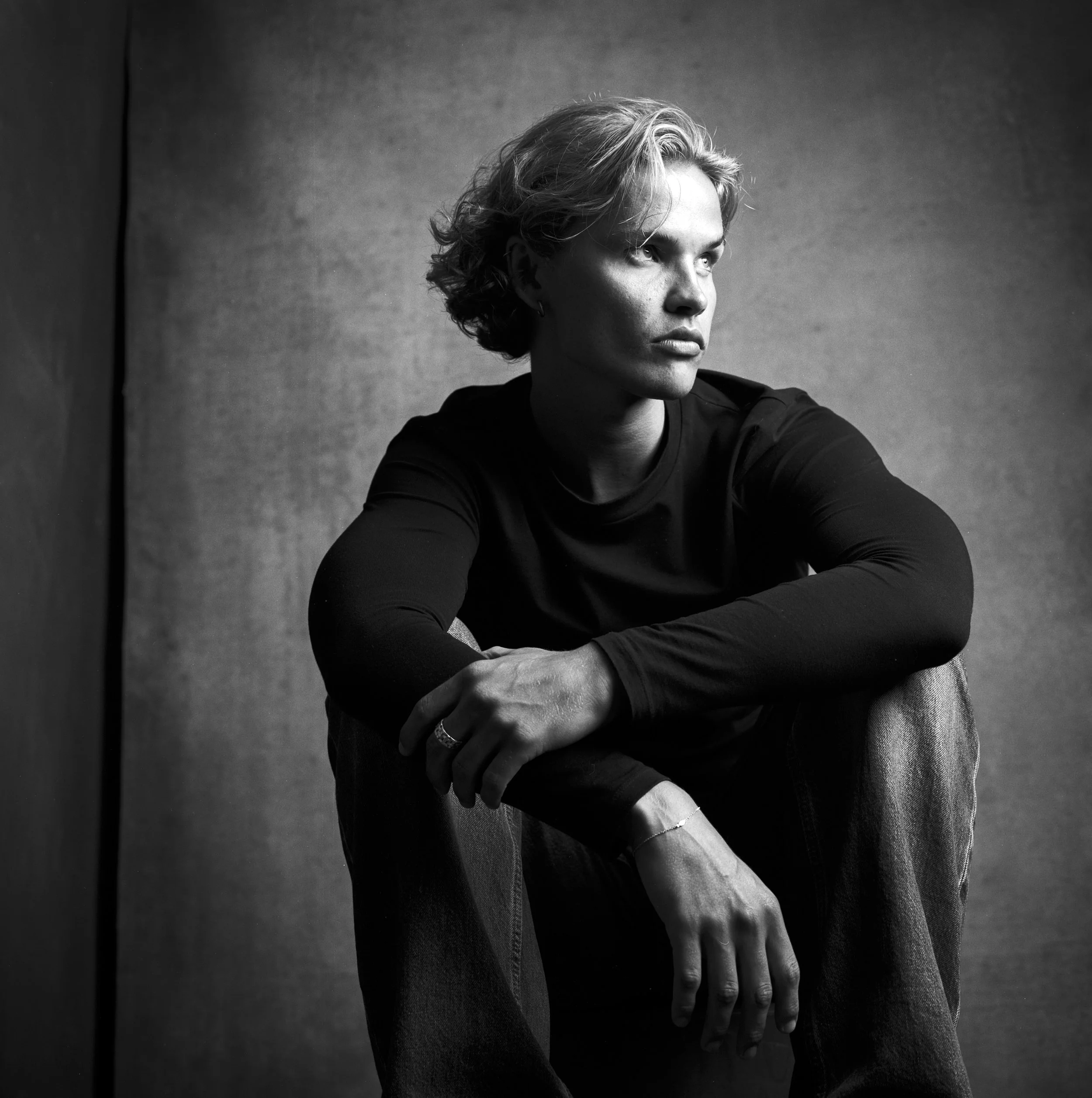 A black-and-white image of a person with short hair and intense eyes sitting and looking to the side, with their arms crossed and a serious expression, against a plain background.