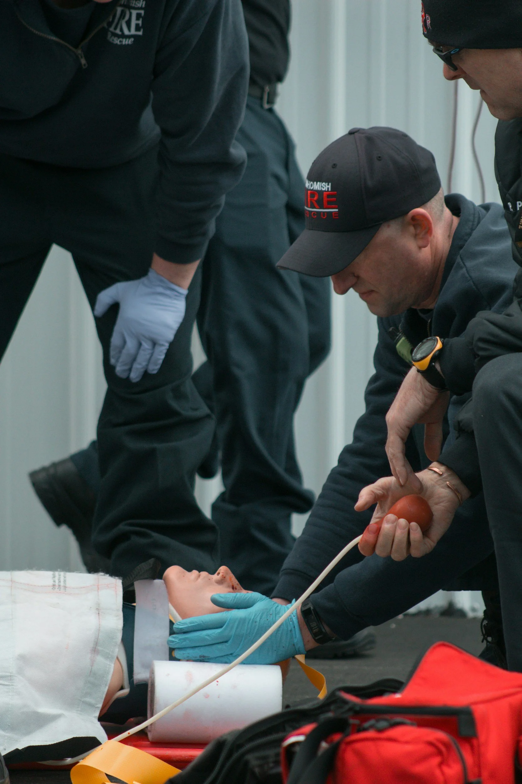 Emergency medical team attends to a patient lying on a cot, with one rescuer checking the patient's pulse and others nearby. The scene appears to be a medical emergency or rescue situation.