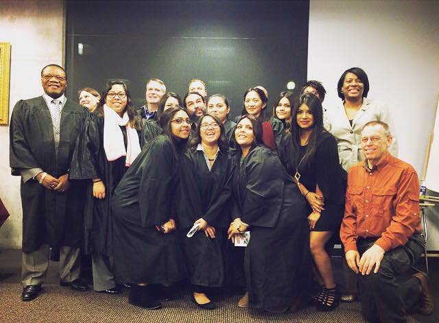 Group of diverse people in graduation gowns and caps posing for a photo in a classroom or conference room.