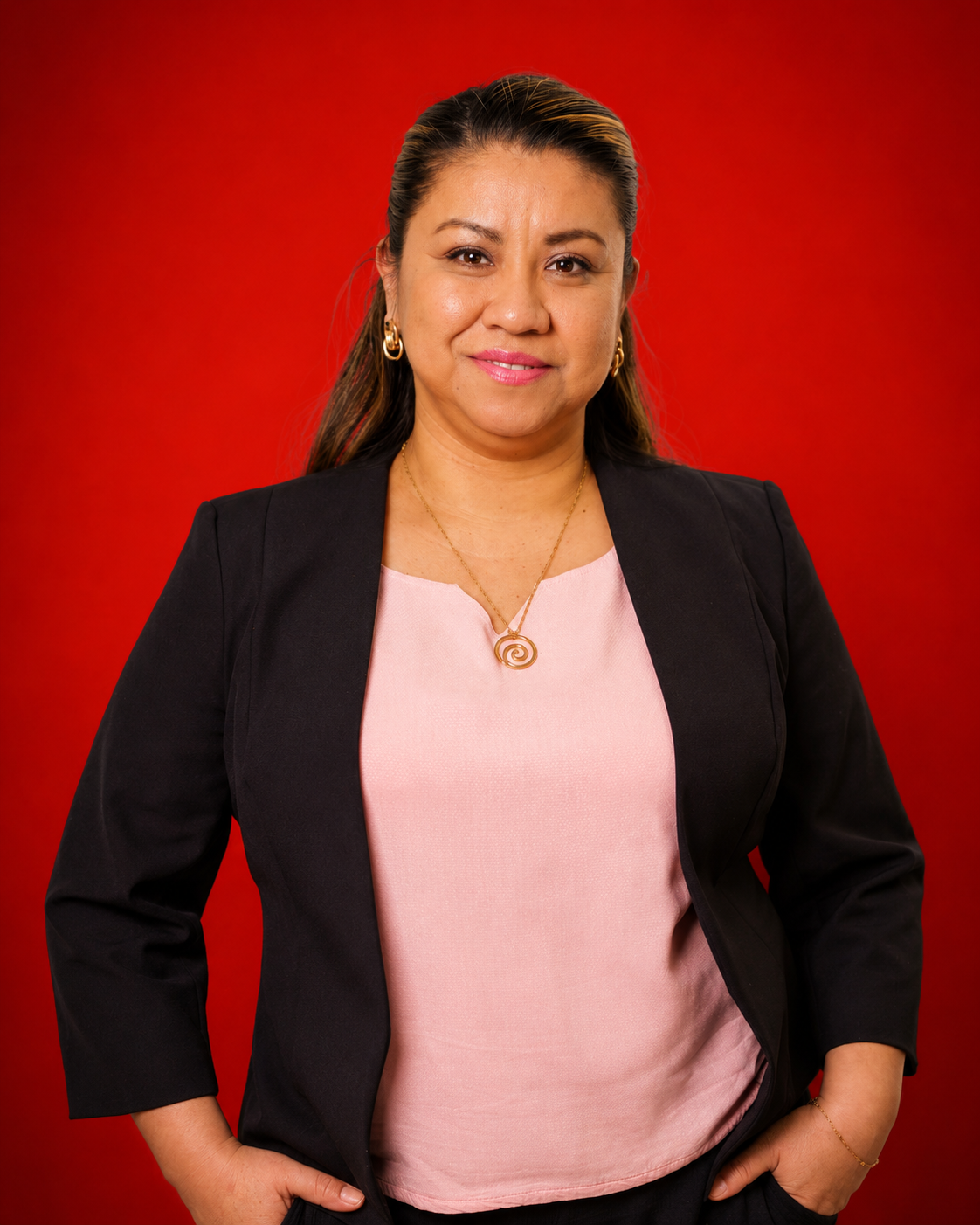A woman with long hair wearing a black blazer over a pink blouse, standing against a red background.