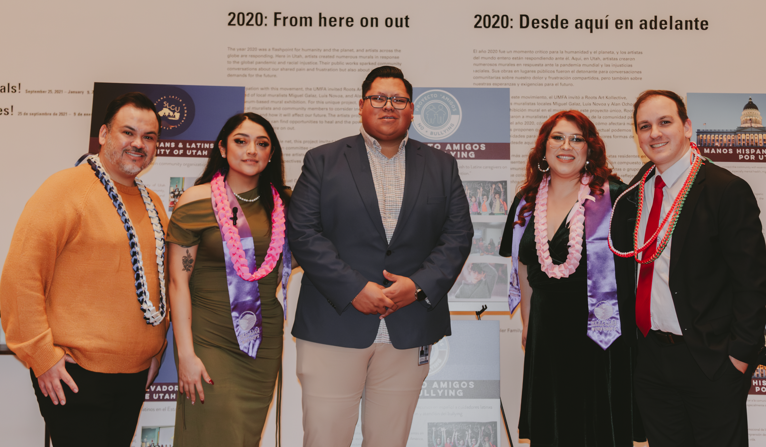 A group of six people standing together indoors in front of informational posters. They are dressed in semi-formal attire with some wearing leis and scarves, and appear to be at a community or cultural event. Most are smiling and looking at the camer