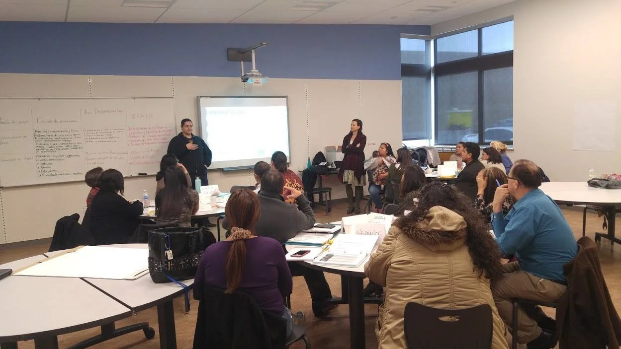 A teacher speaking to a classroom full of adult students in a university or training room, with a whiteboard and a projection screen.