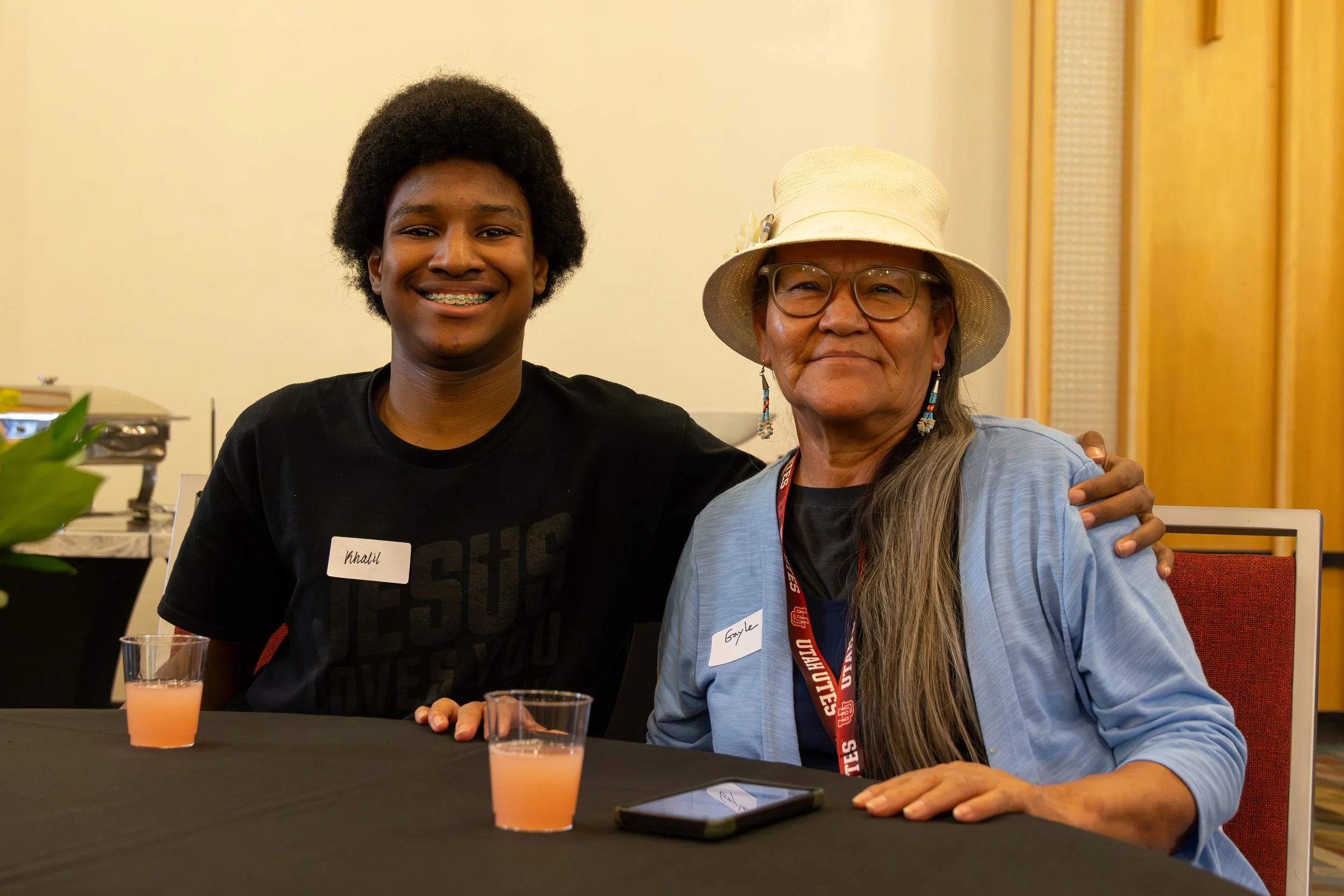 A smiling young man with braces and a woman wearing glasses and a hat, both sitting at a table with drinks, in an indoor setting.