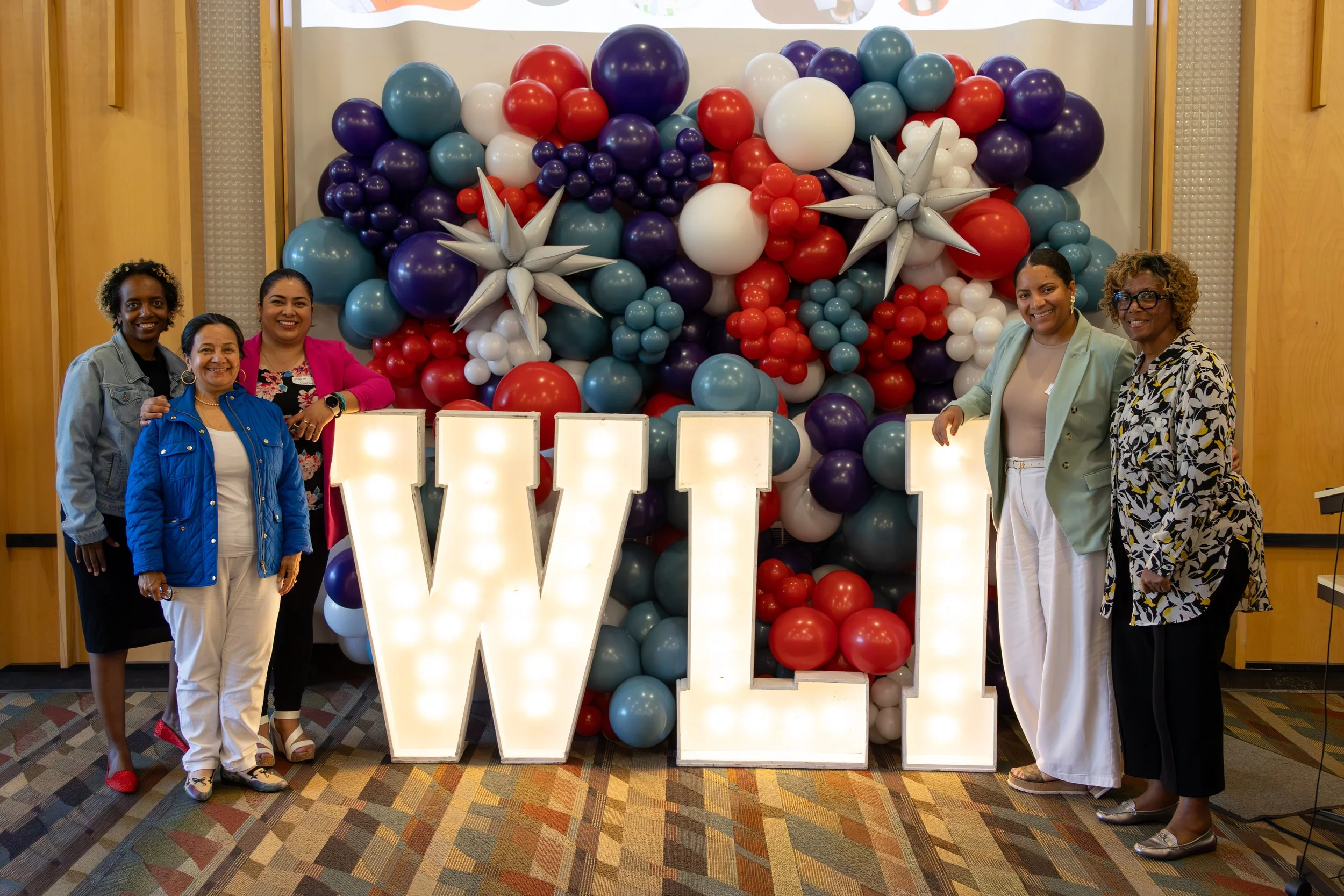 Five women standing in front of a decorative balloon display with the word W on lights. The women are smiling, dressed in various colorful outfits, and are standing indoors on a patterned carpet.