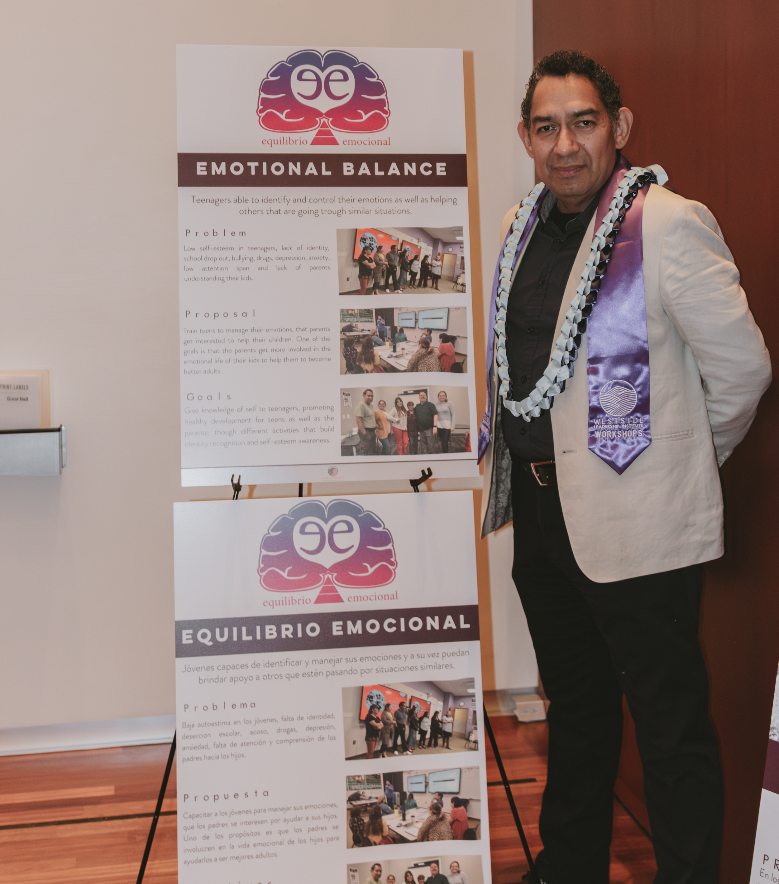 An man dressed in a white blazer with a purple sash and a floral necklace standing next to an informational poster about emotional balance. The poster features a logo with a brain, and headings in Spanish and English, with images of groups of people 