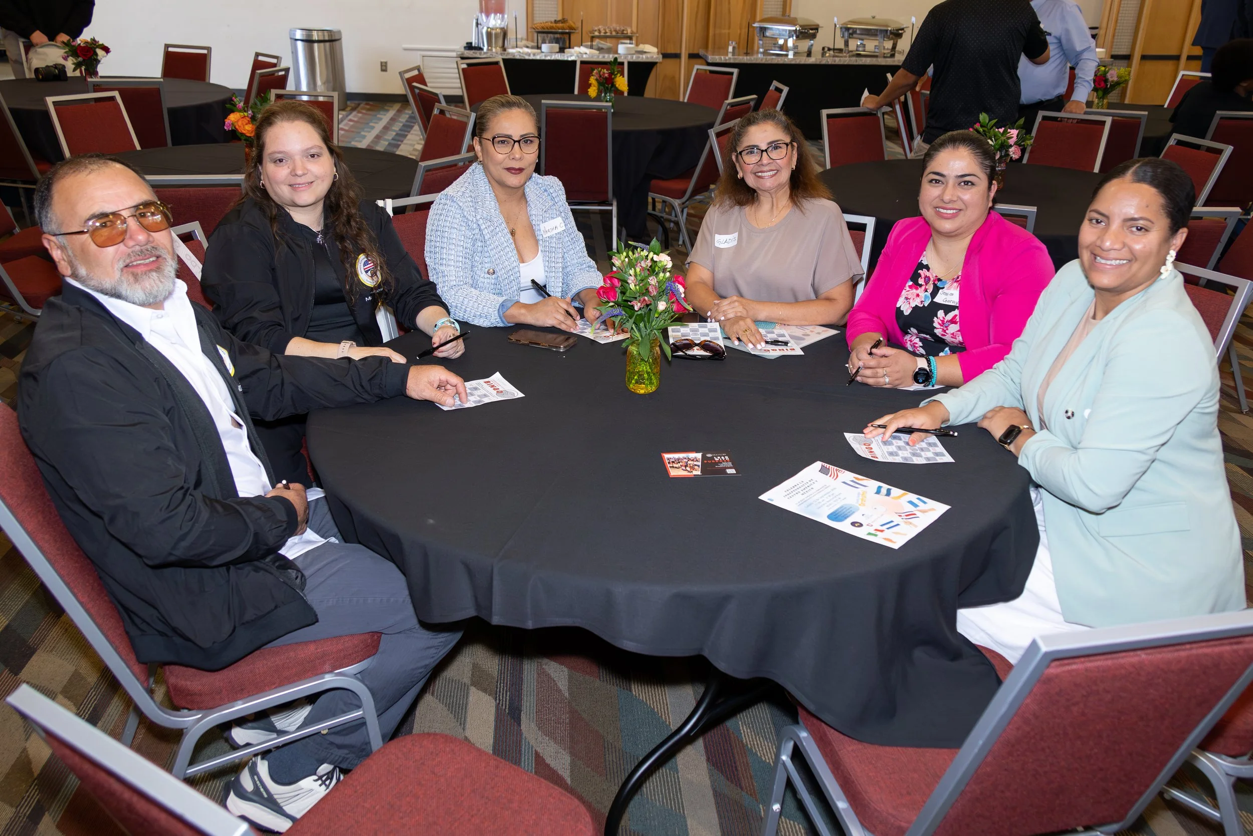 A group of six diverse people sitting around a round black table at a formal event, with a bouquet of pink and green flowers in the center, and brochures and pens on the table. The background shows empty chairs, tables, and a buffet area.