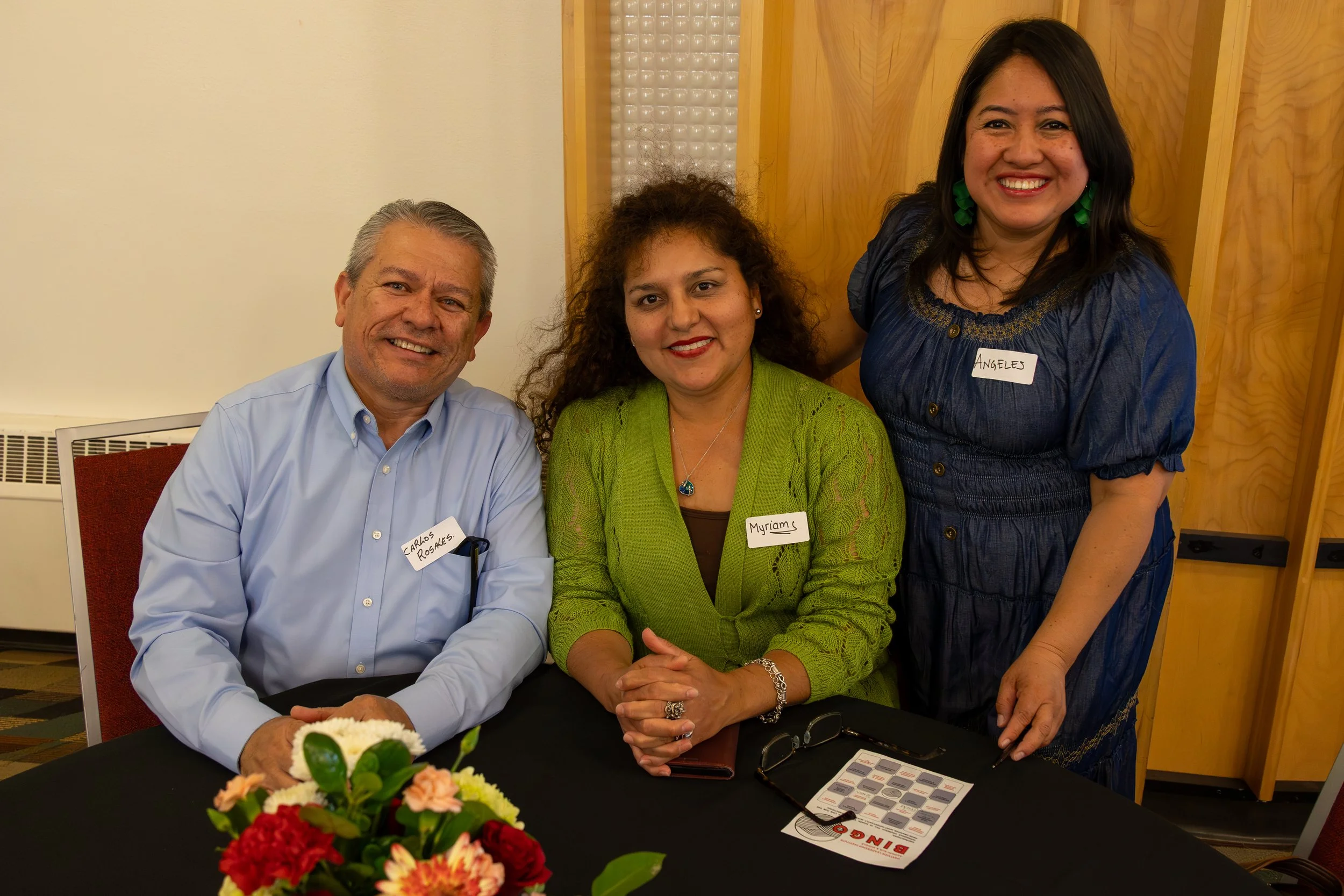 Three people sitting at a table, smiling, with a bouquet of flowers in front. One person standing behind them. The two seated individuals wear name tags that say 'Carlos Rosales' and 'Myriam', and the standing person has a name tag reading 'Ángeles'.