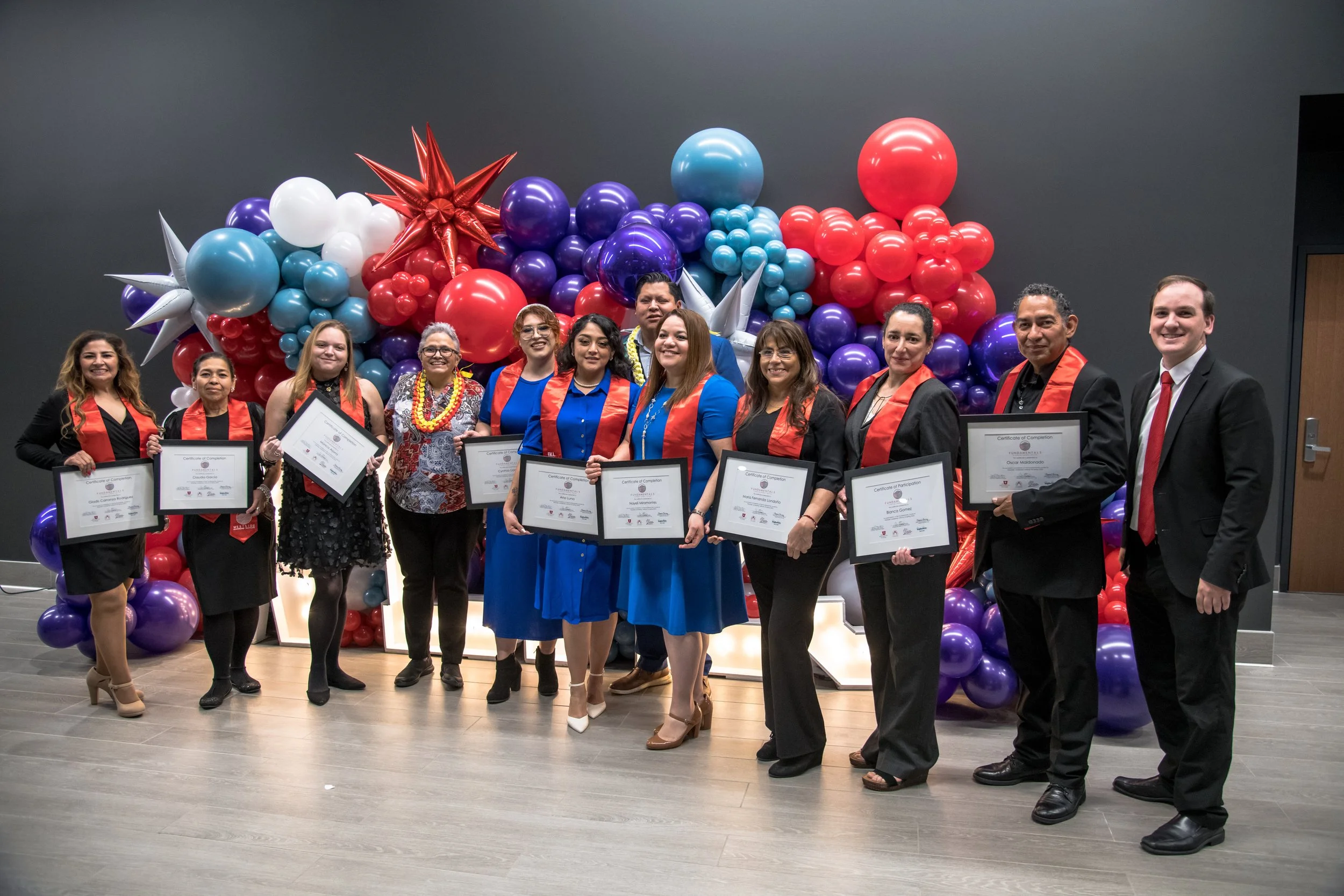 A group of people dressed in formal attire, holding certificates, standing in front of a backdrop of colorful balloons and star-shaped decorations at an indoor celebration or graduation event.