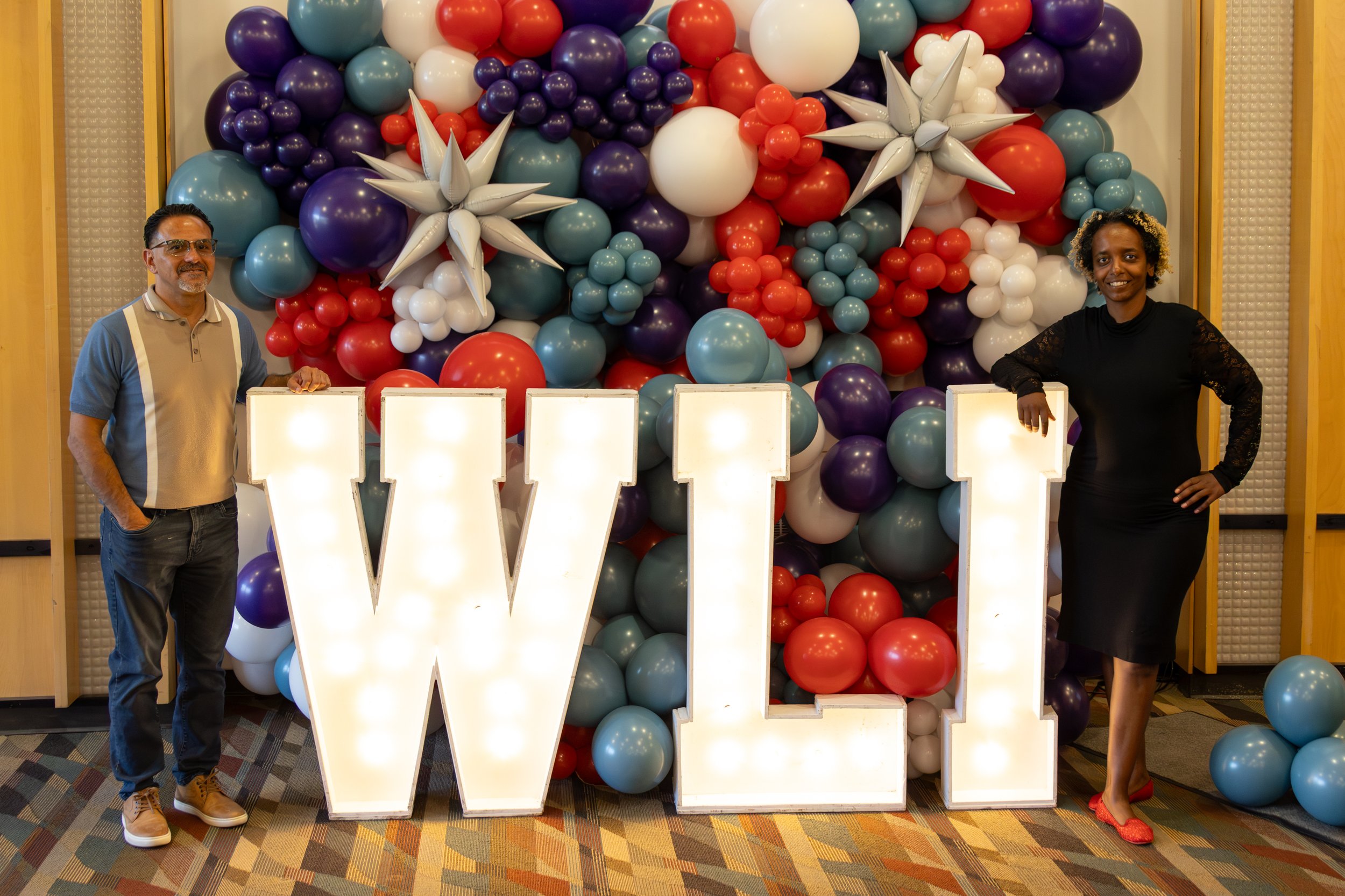 Two people, a man and a woman, standing on either side of a large decorative letter W made of illuminated panels, in front of a colorful balloon wall with balloons in shades of purple, blue, red, and white arranged in clusters and star-shaped balloon