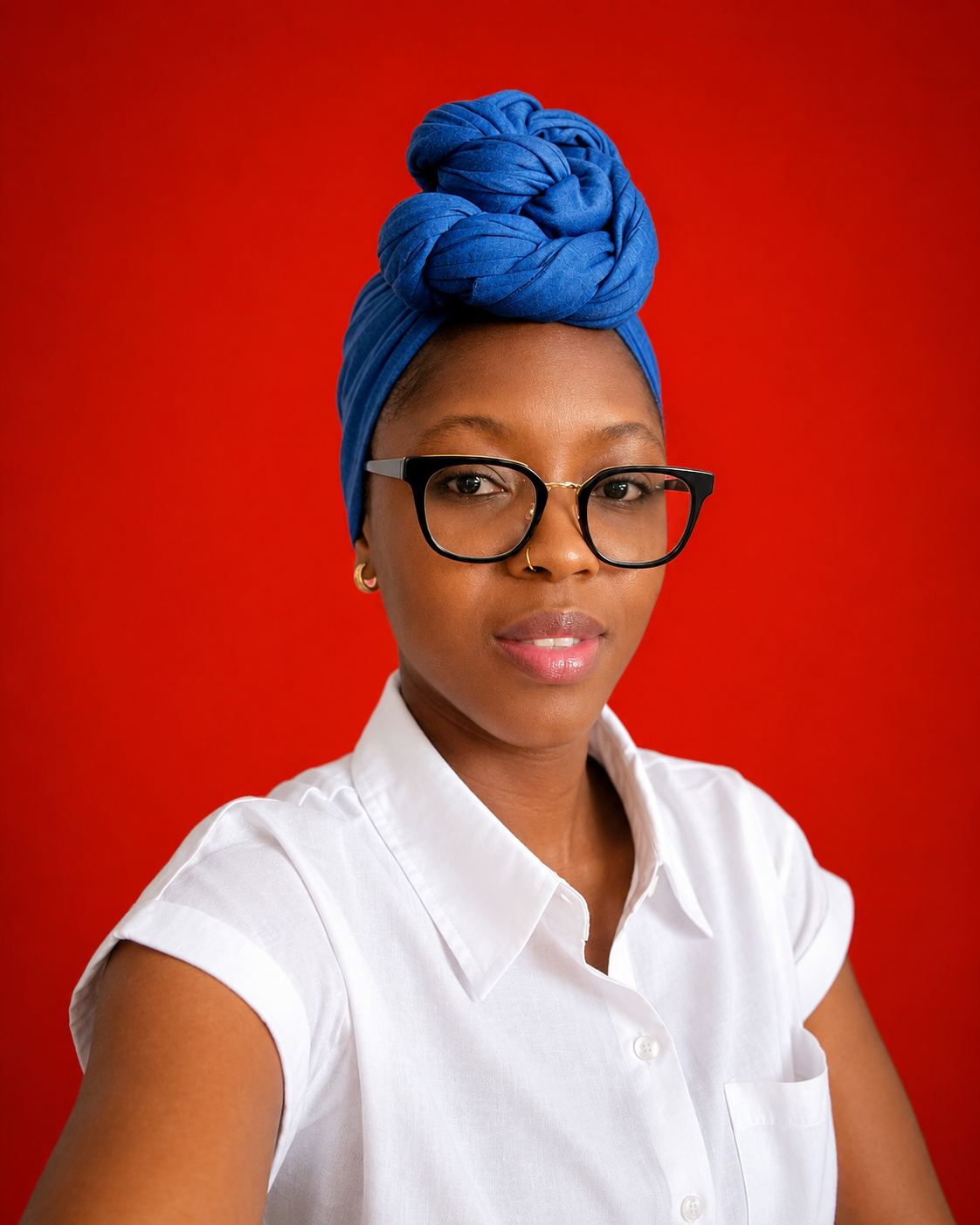 A woman with dark skin wearing glasses, a blue headwrap styled in a knot on top, a white collared shirt, gold earrings, and a nose ring, posing against a red background.