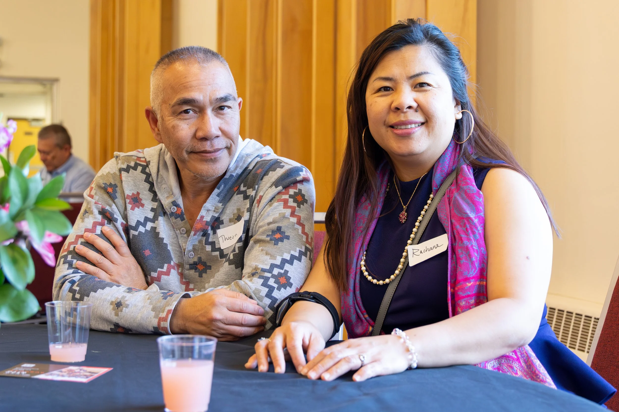 A man and woman sitting at a table with drinks, smiling at the camera, at a social event. The man has short hair and is wearing a patterned shirt. The woman has long hair, wearing hoop earrings, a pearl necklace, and a pink and purple scarf. Both are
