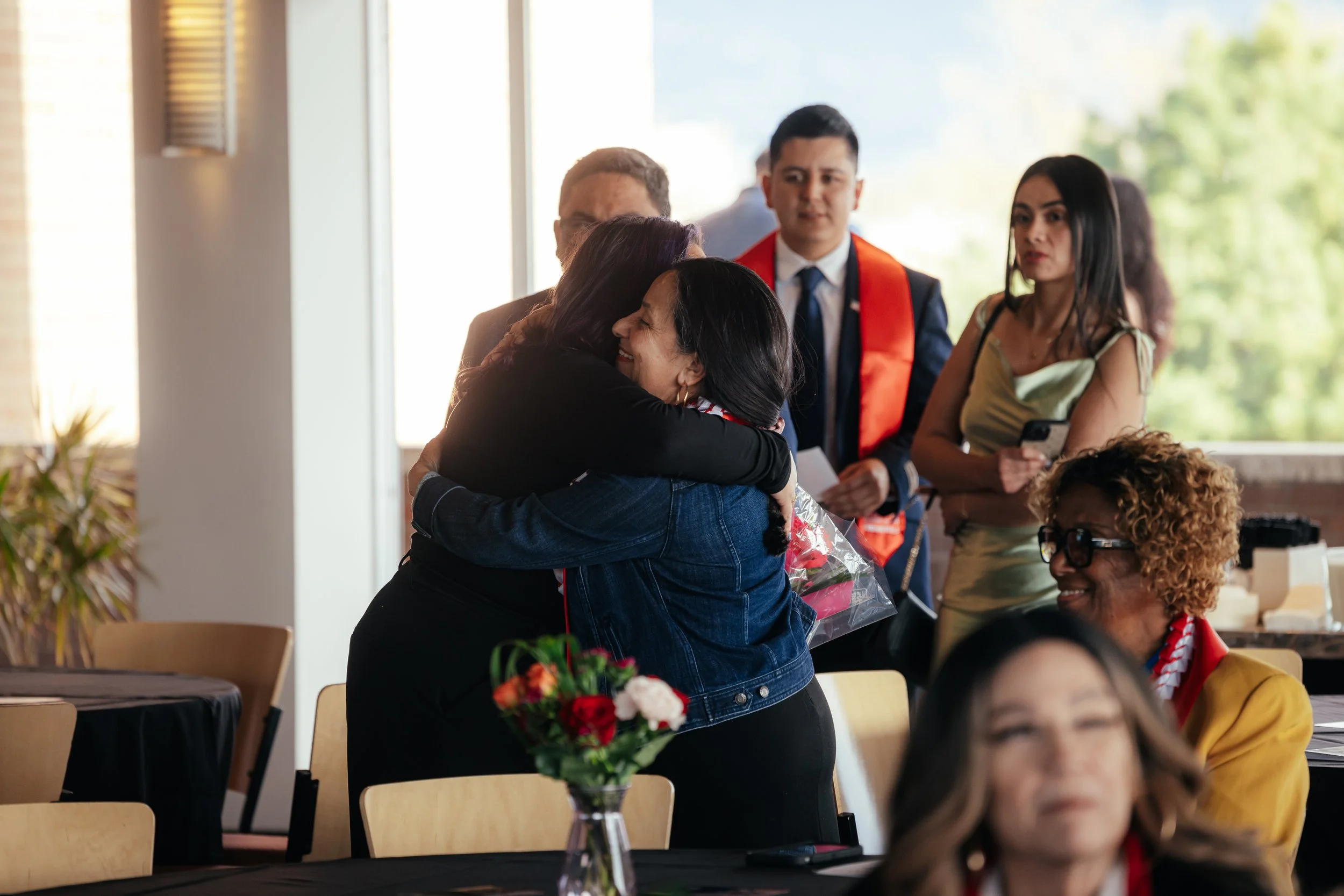 A group of people gathered indoors at a celebration, with two women hugging, one holding a bouquet of flowers. Others are observing, some with expressions of happiness and surprise.