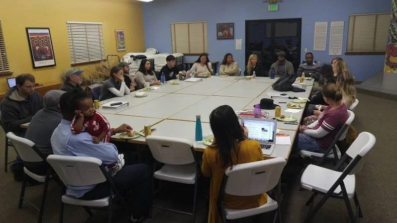 Group of people seated around a large rectangular table in a meeting room, engaging in discussion.