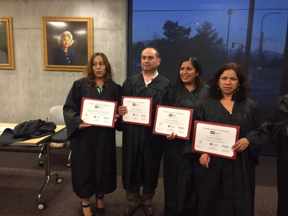 Four people in graduation gowns holding certificates pose for a photo inside a room with artwork on the wall and a large window showing a dark outdoor scene.