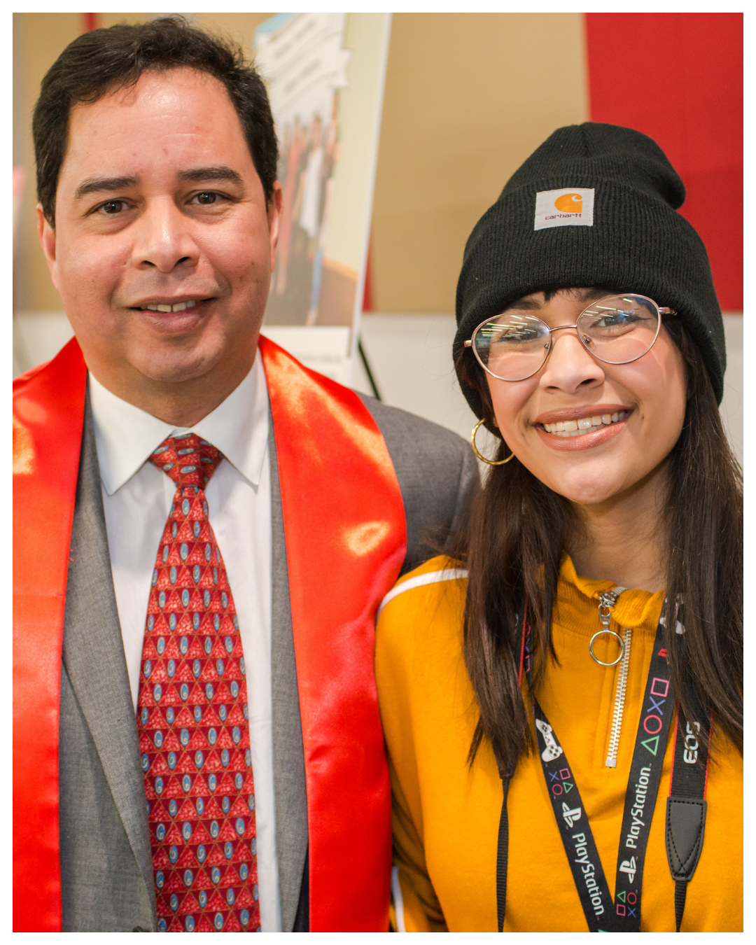 A man in a gray suit and red tie with a red sash standing next to a woman in a yellow jacket, black beanie, and glasses, smiling at the camera.