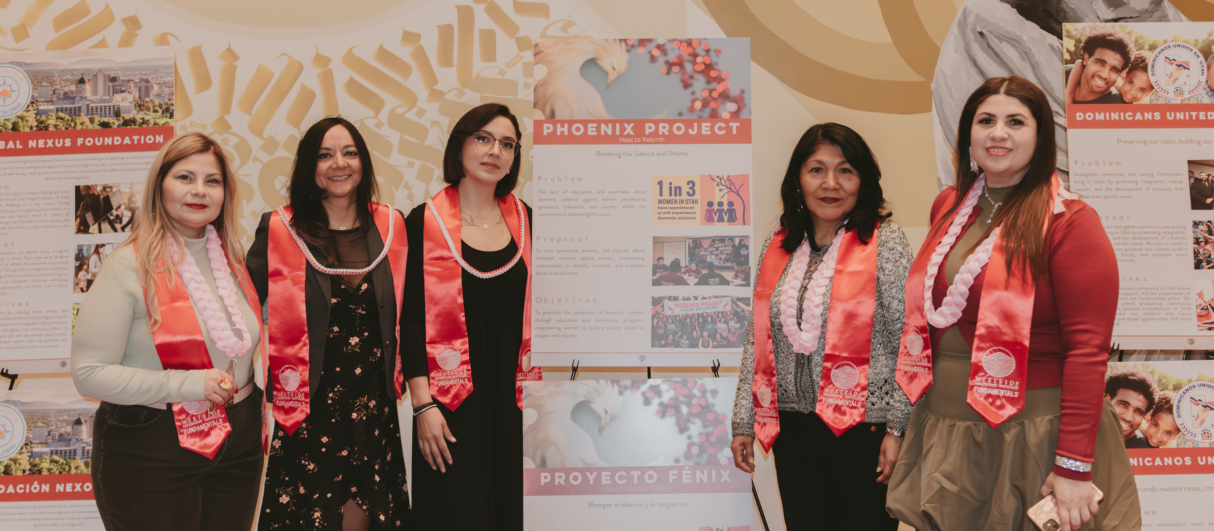 Five women standing in front of display boards at an event, all wearing red sashes with white ribbons, smiling, in a professional or community event setting.