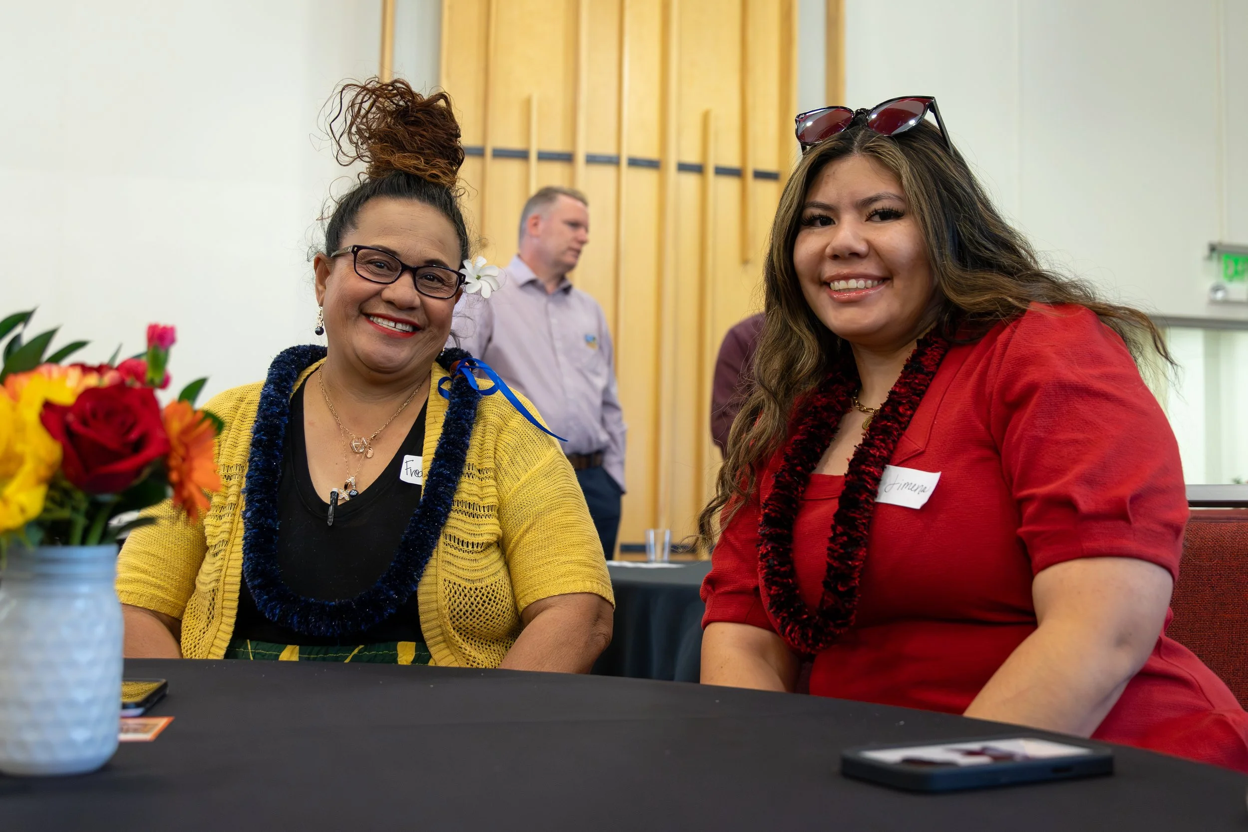 Two women sitting at a table, smiling and looking at the camera. The woman on the left is wearing glasses, a yellow cardigan, and has curly hair tied up. The woman on the right has long brown hair, sunglasses on her head, and is wearing a red outfit.