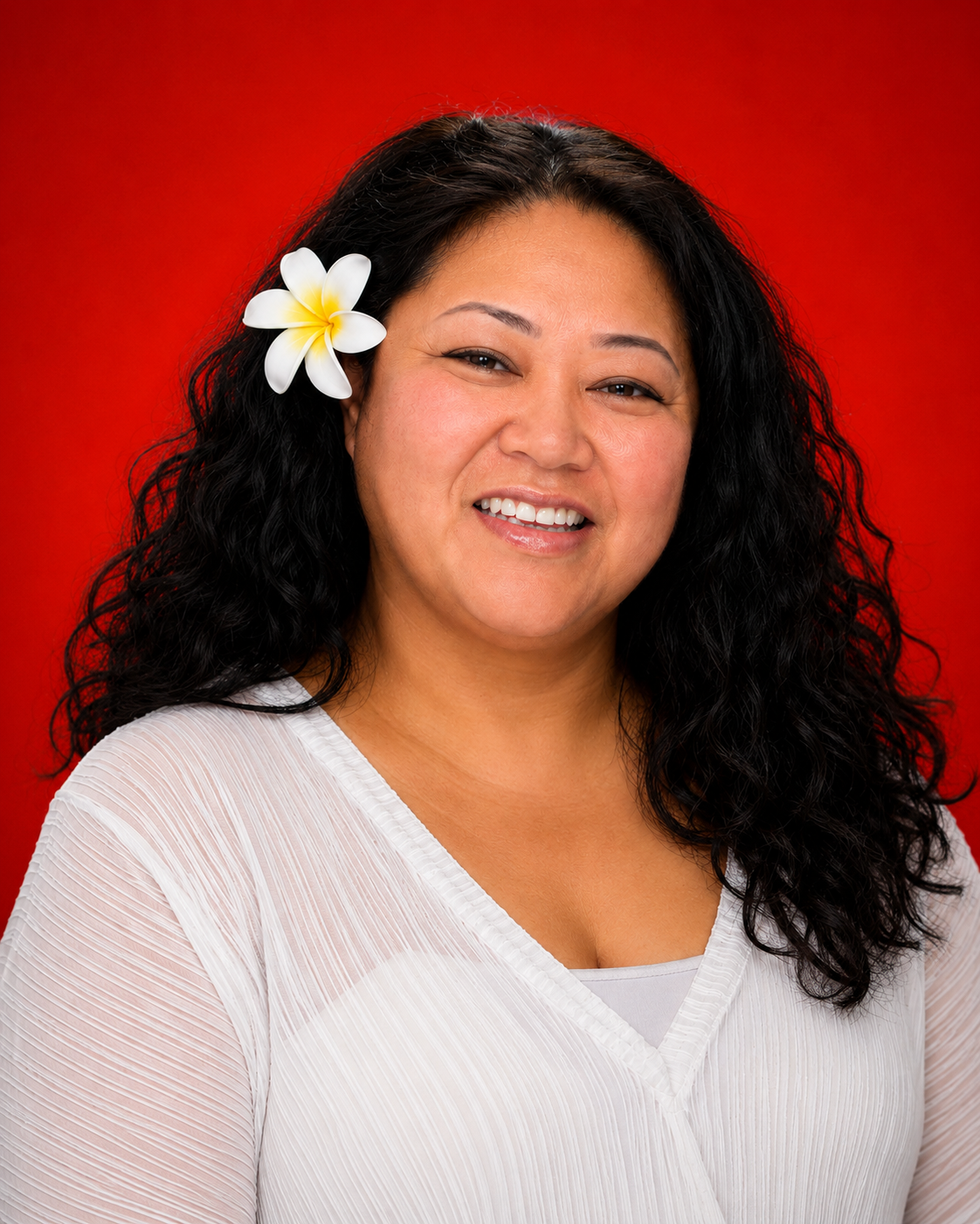 Smiling woman with long curly black hair, a white flower in her hair, wearing a white top, against a red background.