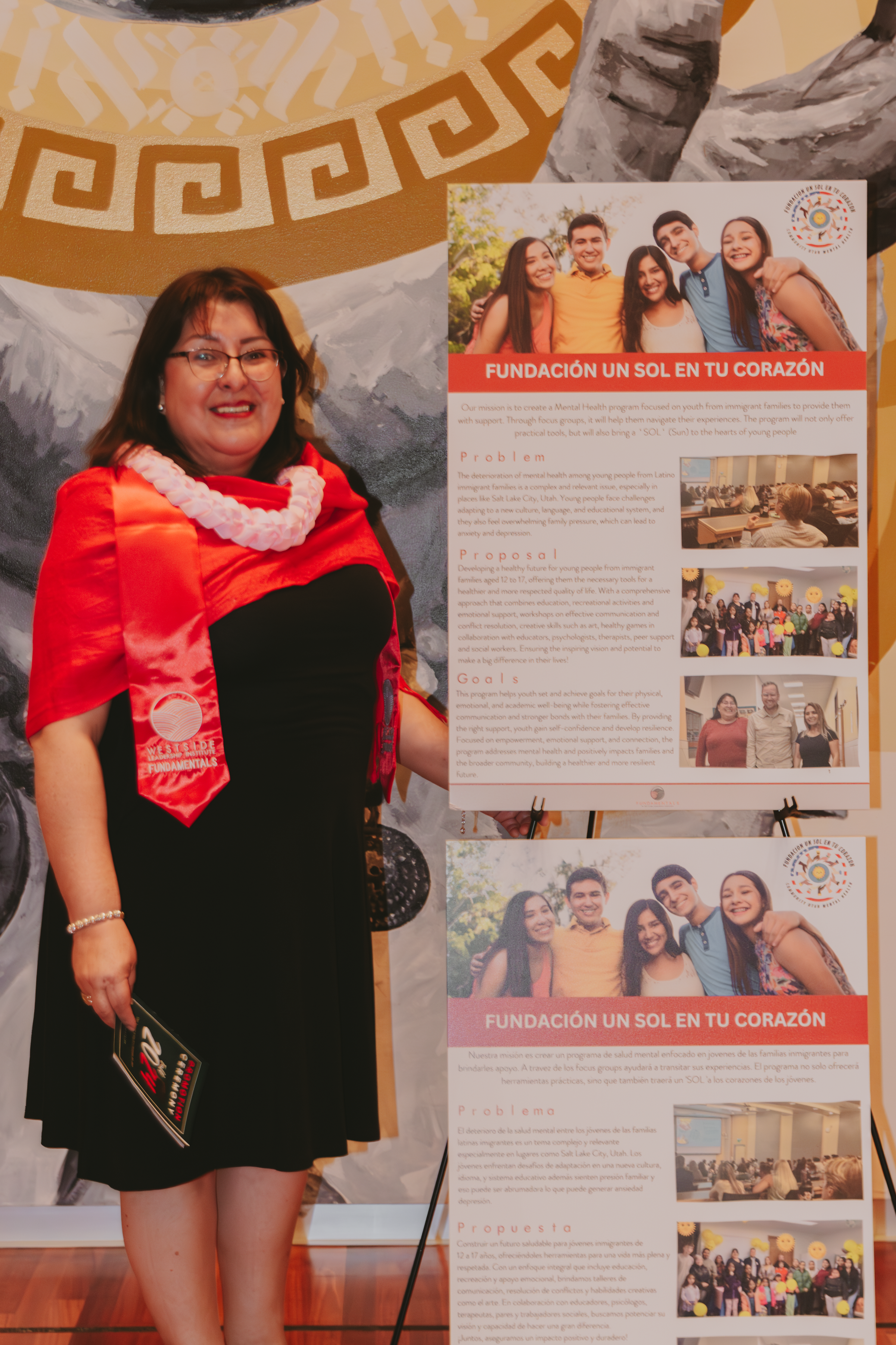 A woman with glasses, wearing a red shawl and black dress, standing next to a poster about a mental health foundation for immigrant youth, with a mural background and a mountain scene.