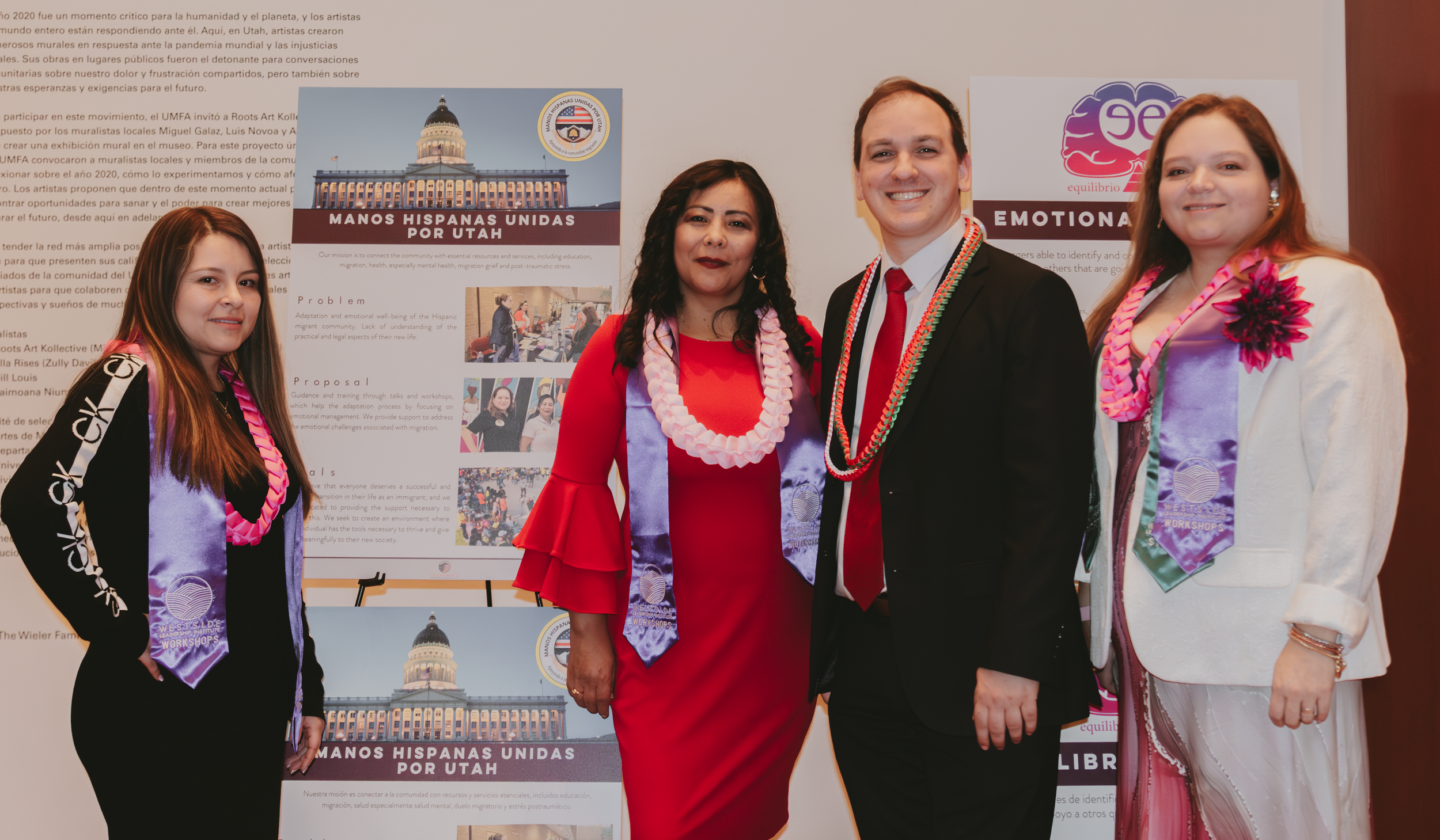 Four people dressed formally at a cultural event, standing in front of informational posters, wearing medals and leis.