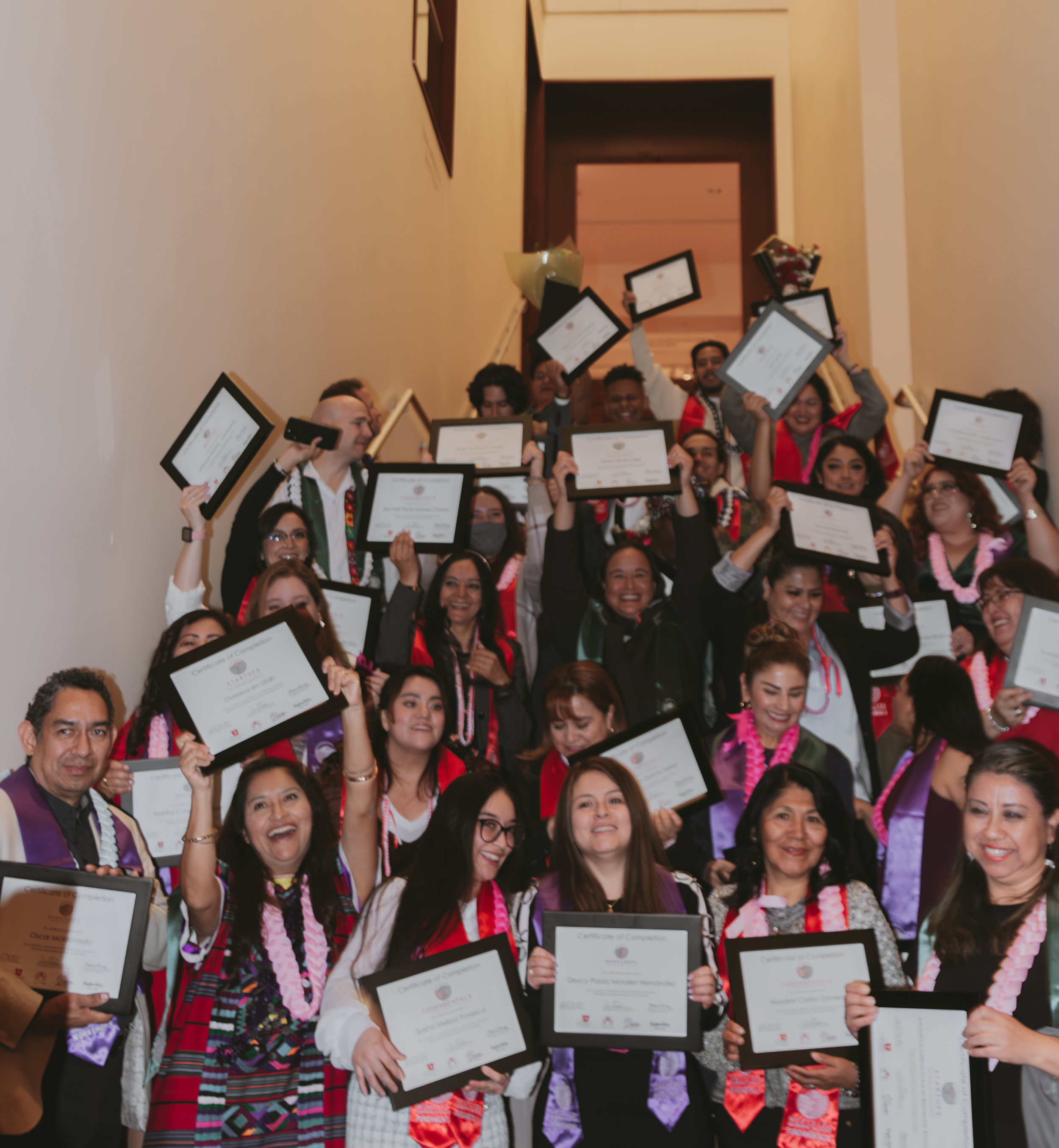 Group of diverse people at a graduation ceremony holding diplomas, some with leis, smiling and celebrating indoors.