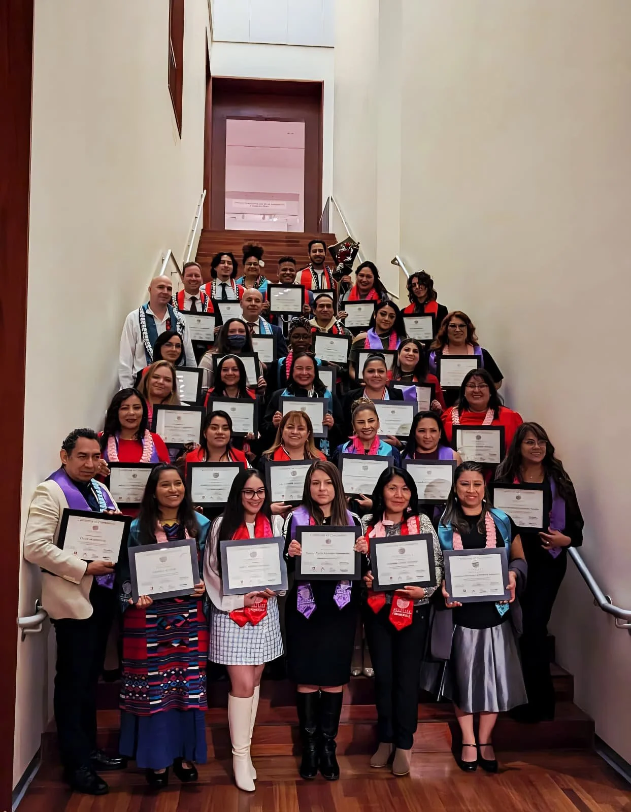 A large group of people pose for a photo on a staircase indoors, holding diplomas and wearing colorful academic regalia, celebrating an achievement.