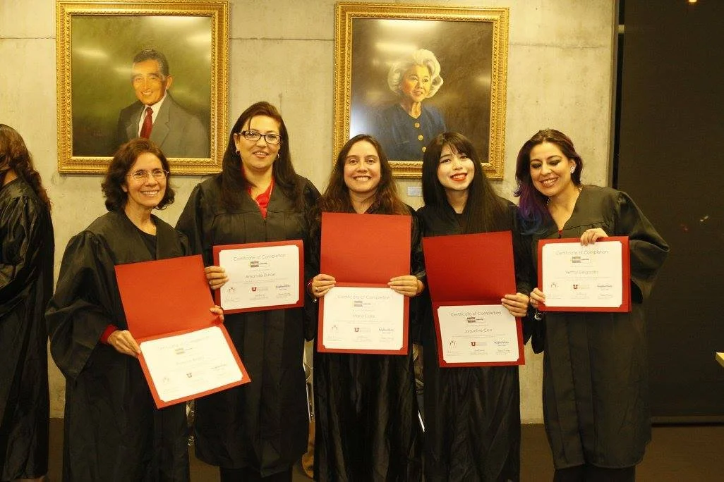 Five women in black graduation gowns holding certificates, standing in front of portrait paintings on a concrete wall.