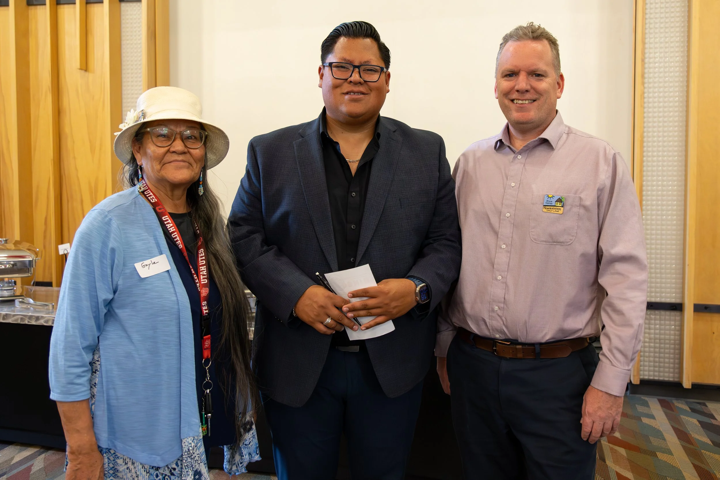 Three people standing together, smiling, indoors with wooden wall panels. The woman on the left wears glasses, a hat, a light blue jacket, and has a Utah Utes lanyard. The man in the middle wears glasses, a dark suit with an open-collared shirt, hold