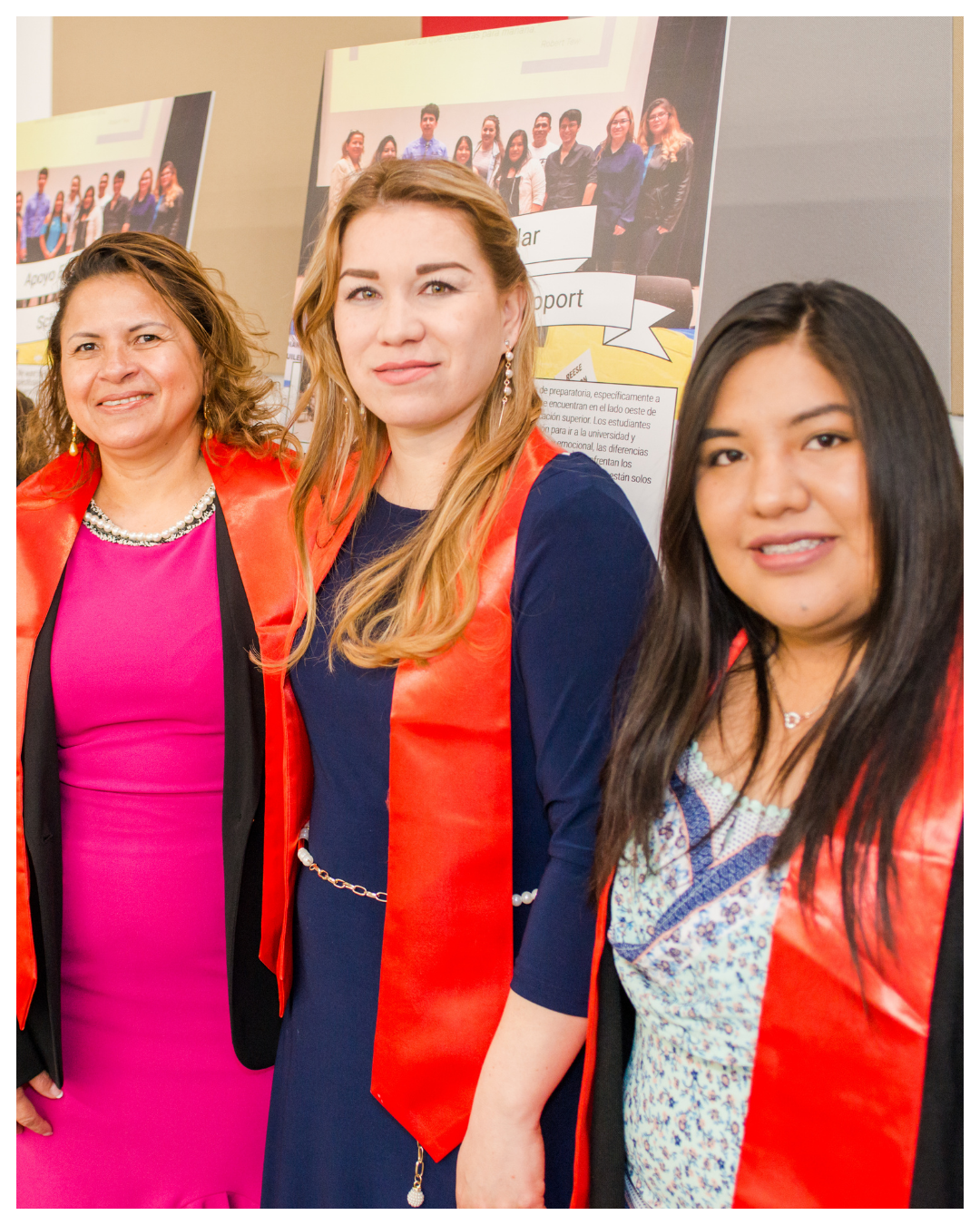 Group of women at a graduation or academic event, wearing red sashes over their formal attire, standing in front of presentation posters.