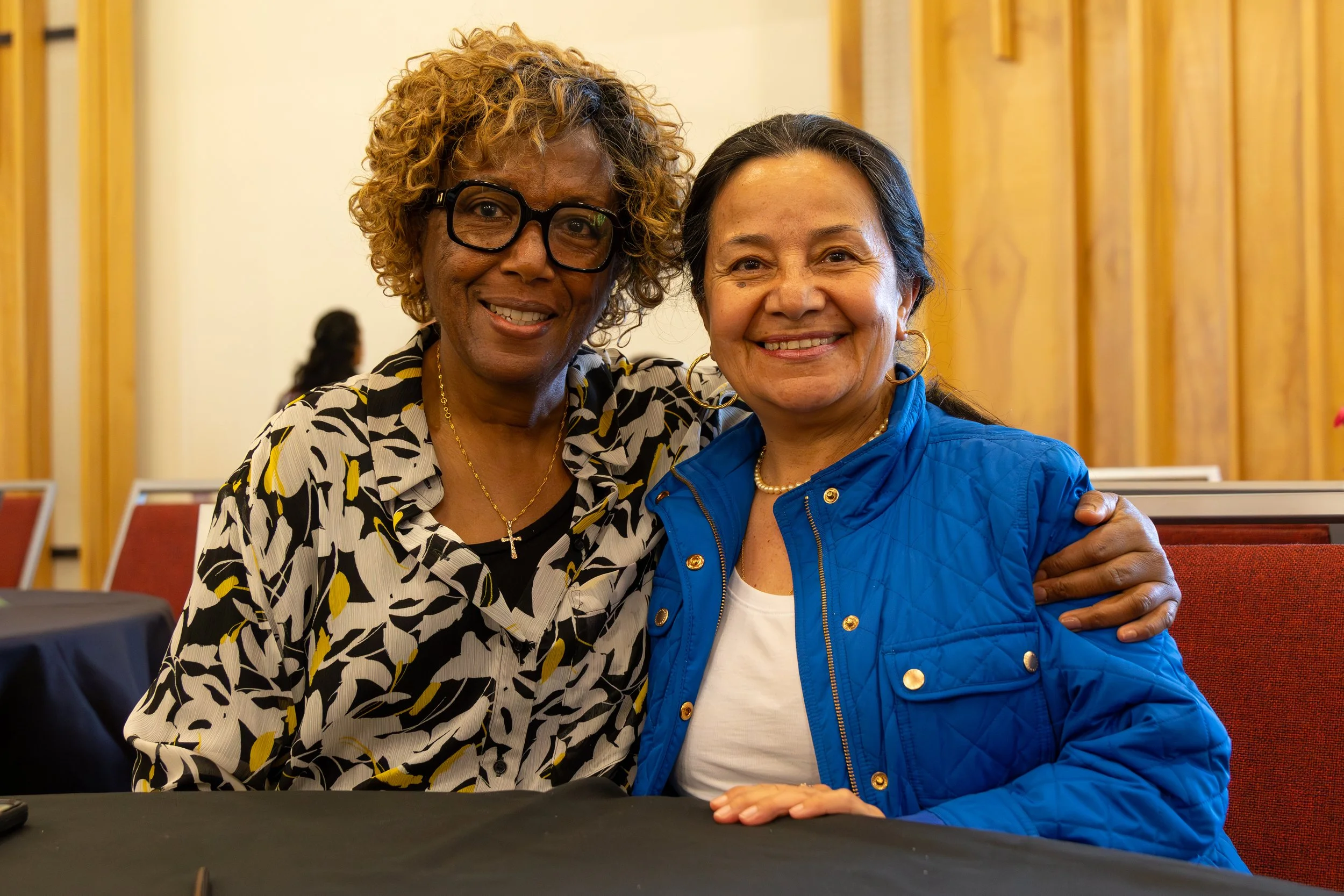 Two women smiling and hugging in an indoor setting, one wearing a patterned blouse with glasses and a cross necklace, the other in a blue jacket and white shirt, with wooden paneling in the background.
