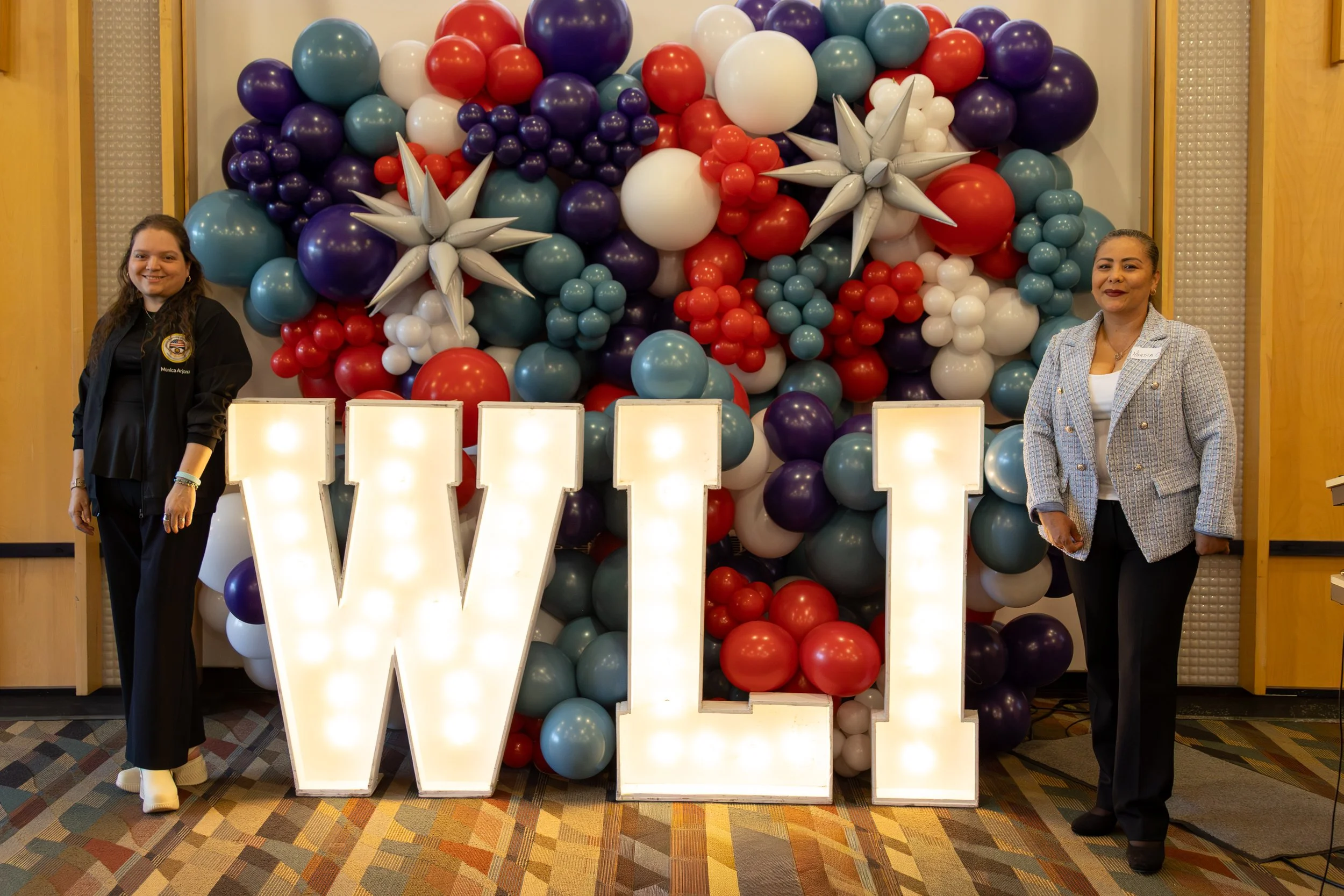 Two women stand on either side of a large decorative balloon display with the letters W and E lit up in front, suggesting the word 'WE'. The balloon arrangement includes balloons in shades of purple, blue, white, red, and teal, with some white star-s