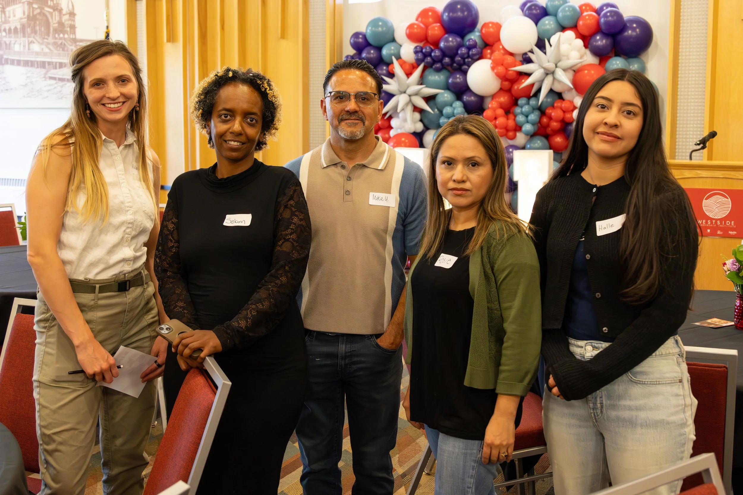 Group of five diverse individuals standing indoors, smiling, with colorful balloon art in the background.