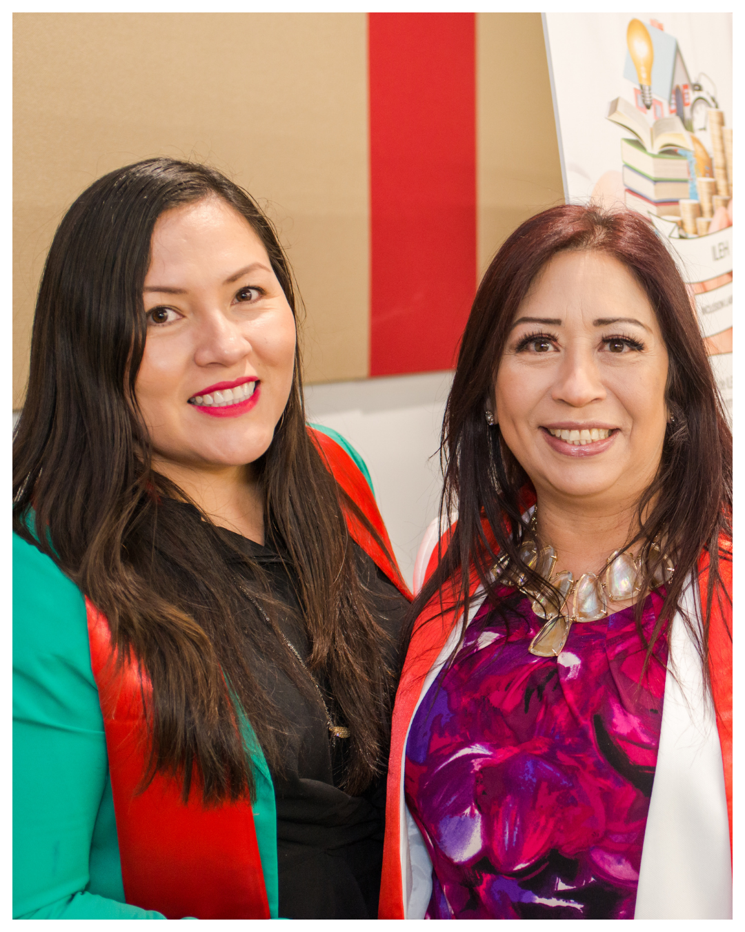 Two women smiling, standing indoors near a colorful wall and a poster with books and a lightbulb illustration.