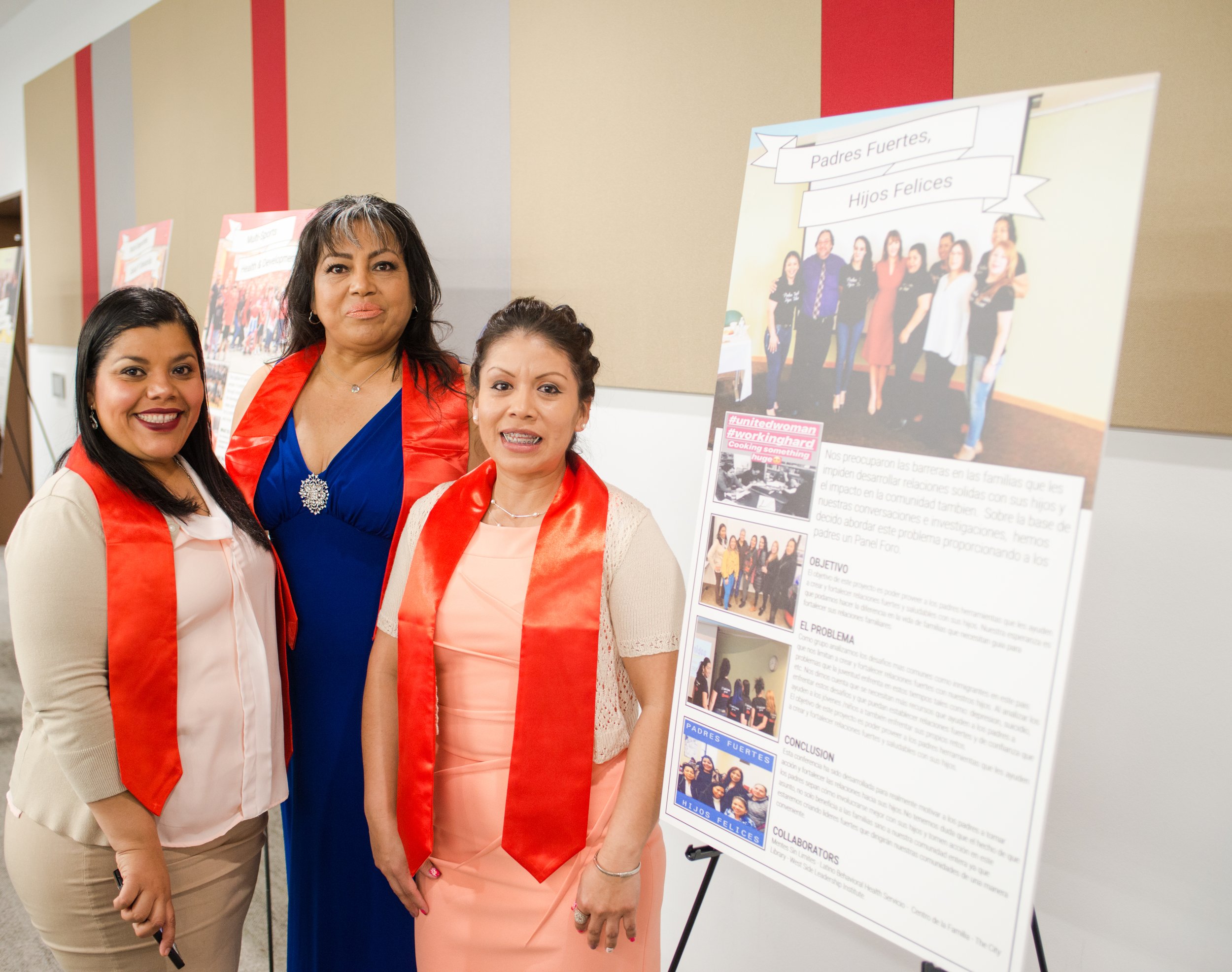 Three women standing next to a large research poster at an indoor event, smiling for the photo. The poster features images and text in Spanish about community relationships and family strength.