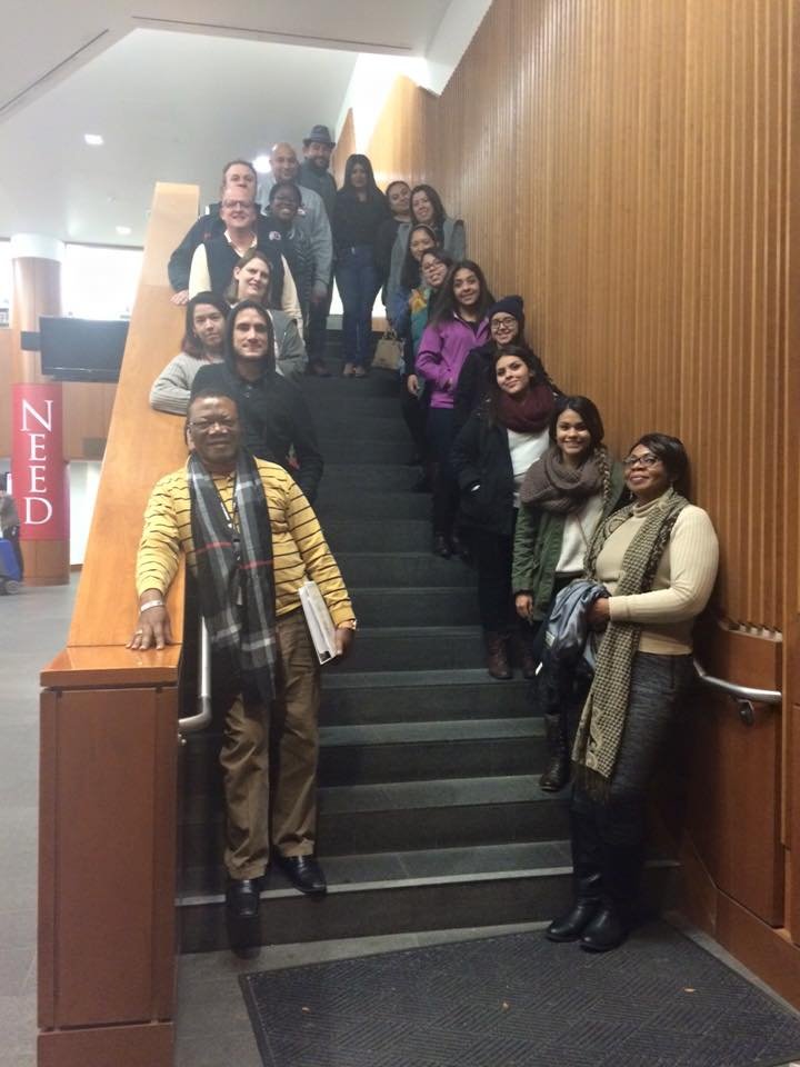 A group of people standing on a staircase in an indoor setting, posing for a photo. The staircase has a wooden handrail and walls with vertical wood paneling.