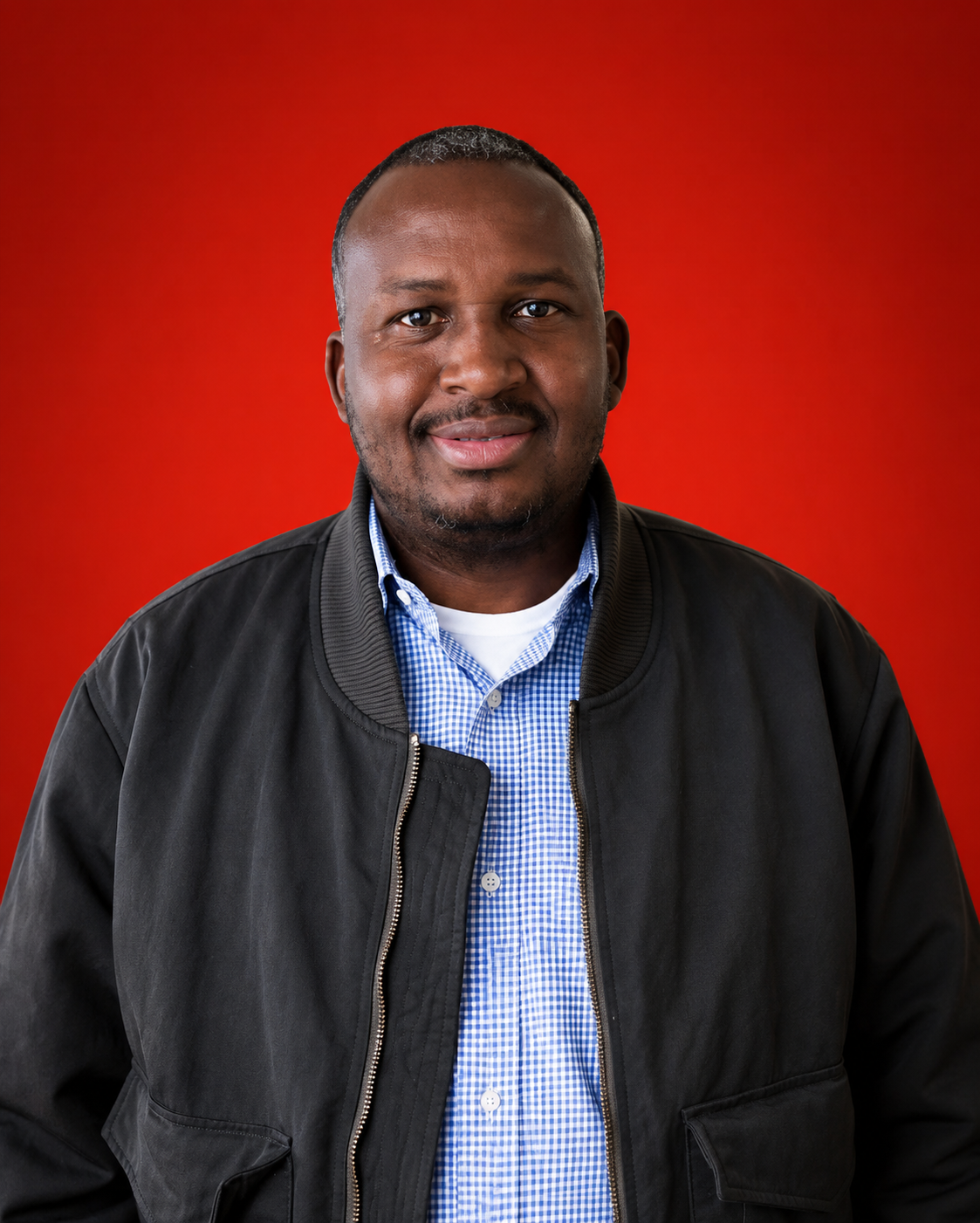 Portrait of a man with short hair and a beard, wearing a black jacket over a blue and white checkered shirt, against a red background.