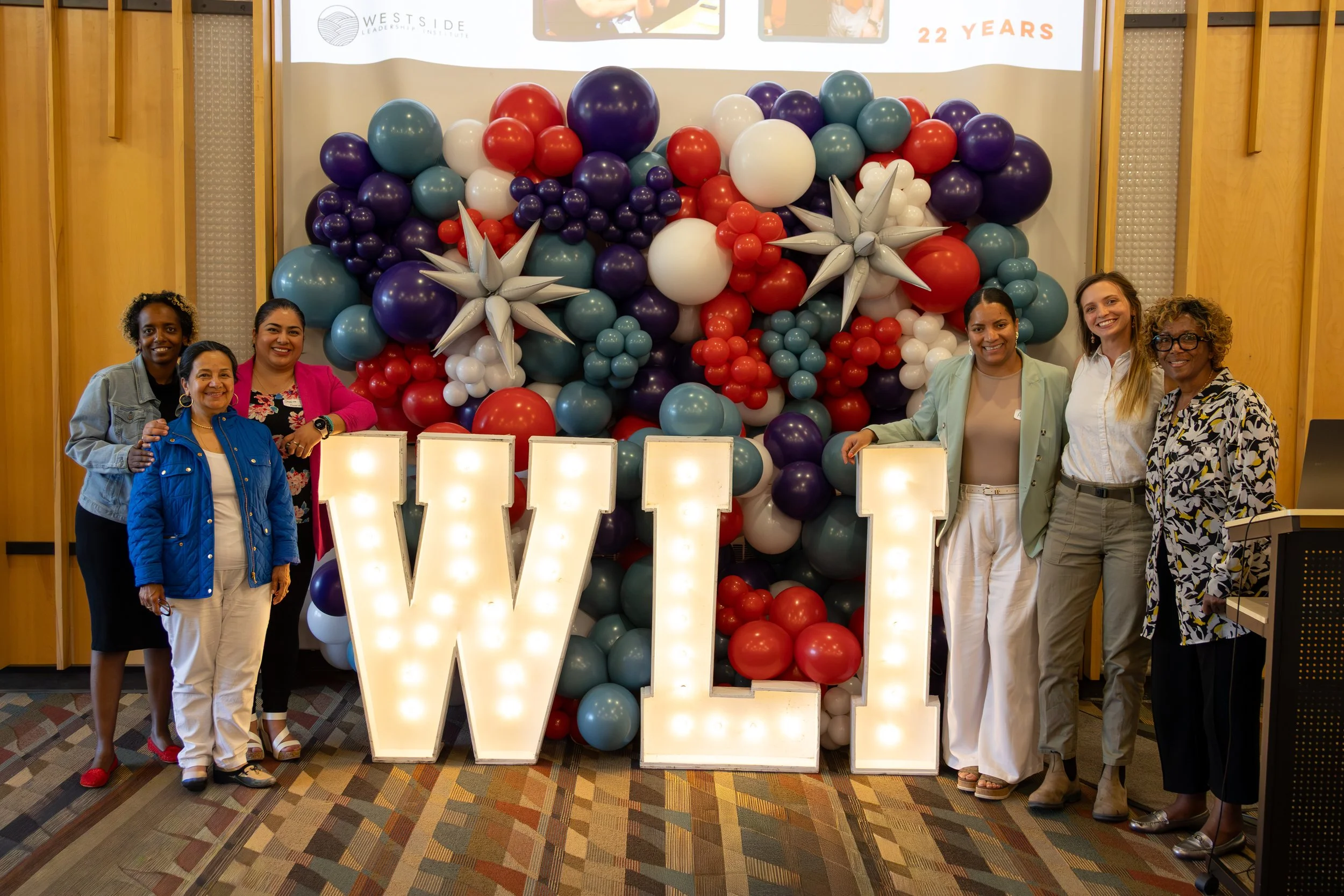 Group of six women standing in front of a colorful balloon display with illuminated letters spelling 'W'. The women are smiling and dressed in business casual attire, with a large screen and wooden walls in the background.