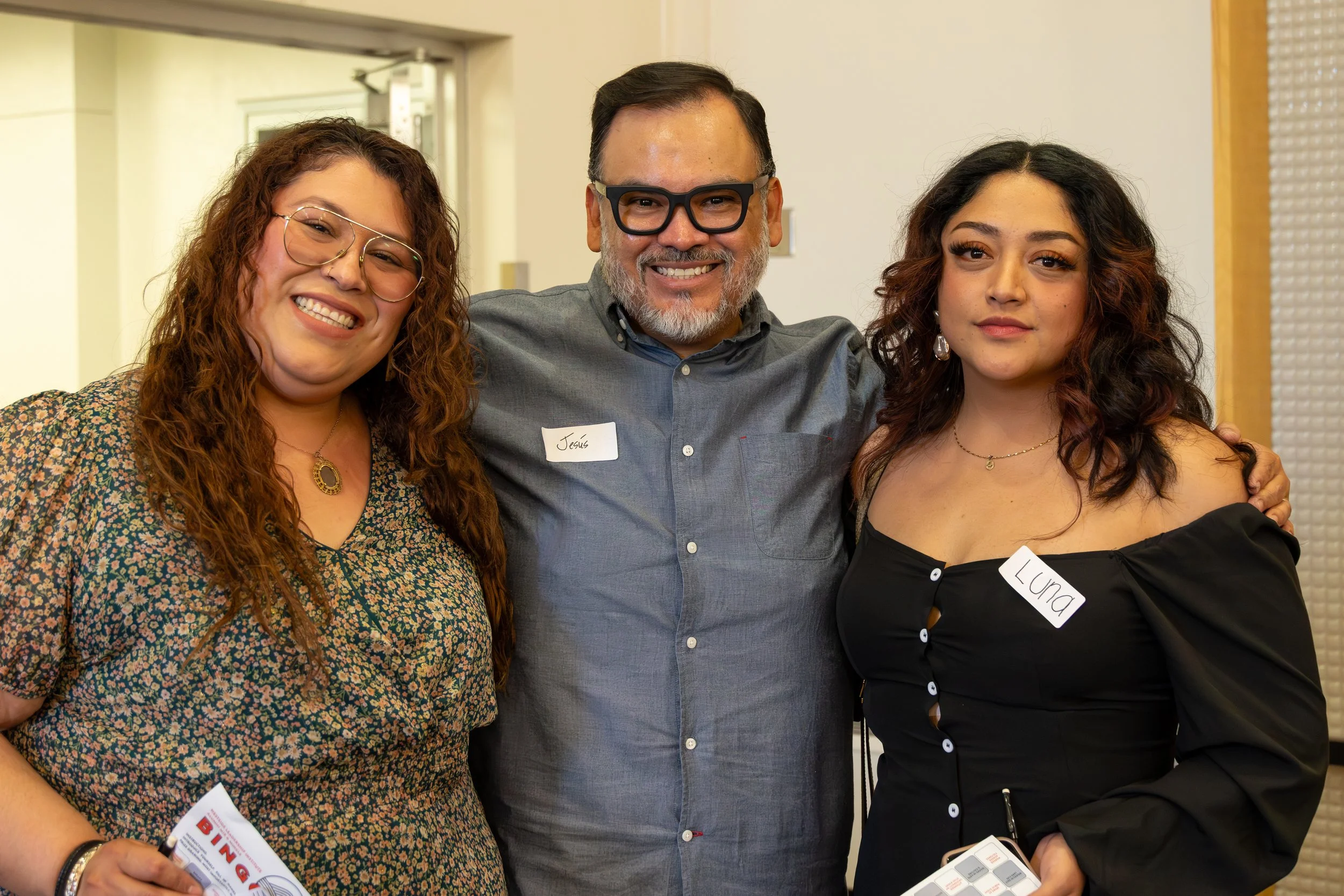 Three people standing close together, smiling at the camera, in an indoor setting. The woman on the left has red curly hair, glasses, and a floral dress. The man in the middle is wearing glasses, a grey shirt with a name tag that reads 'Jesús'. The w