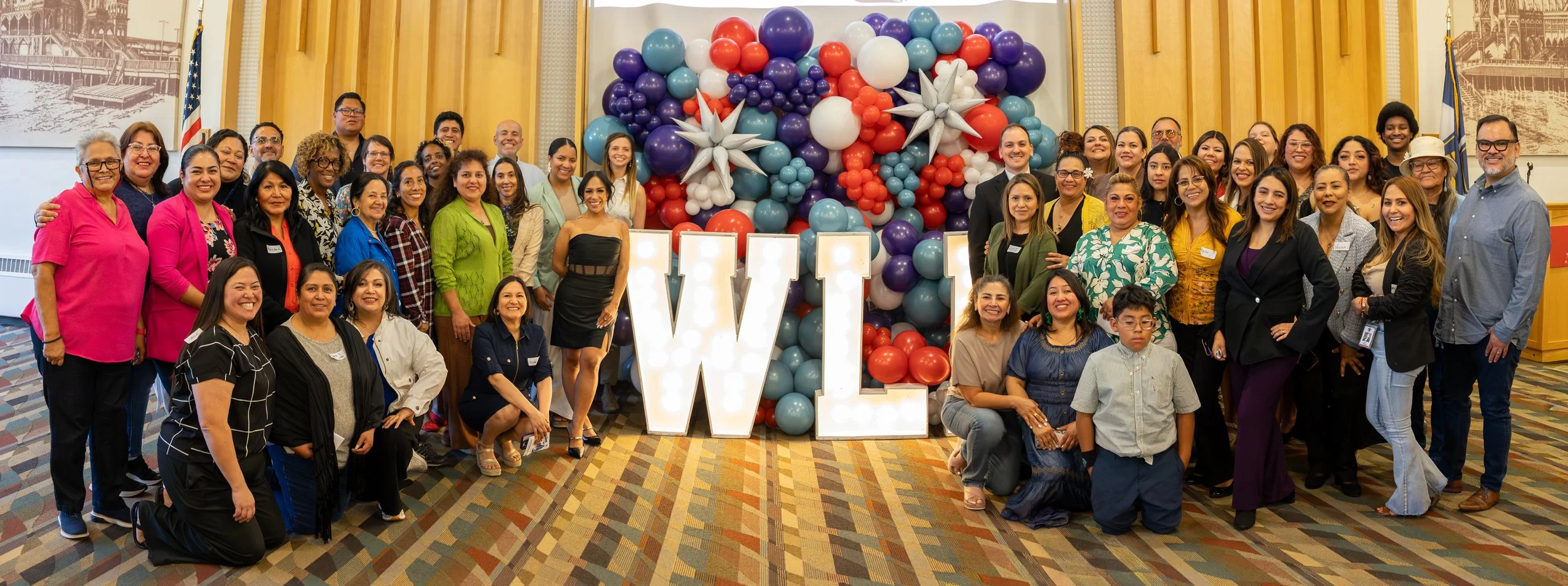 Group of diverse people gathered at an event with a large decorative balloon backdrop, featuring a mix of colors and big illuminated 'W' letters in front.