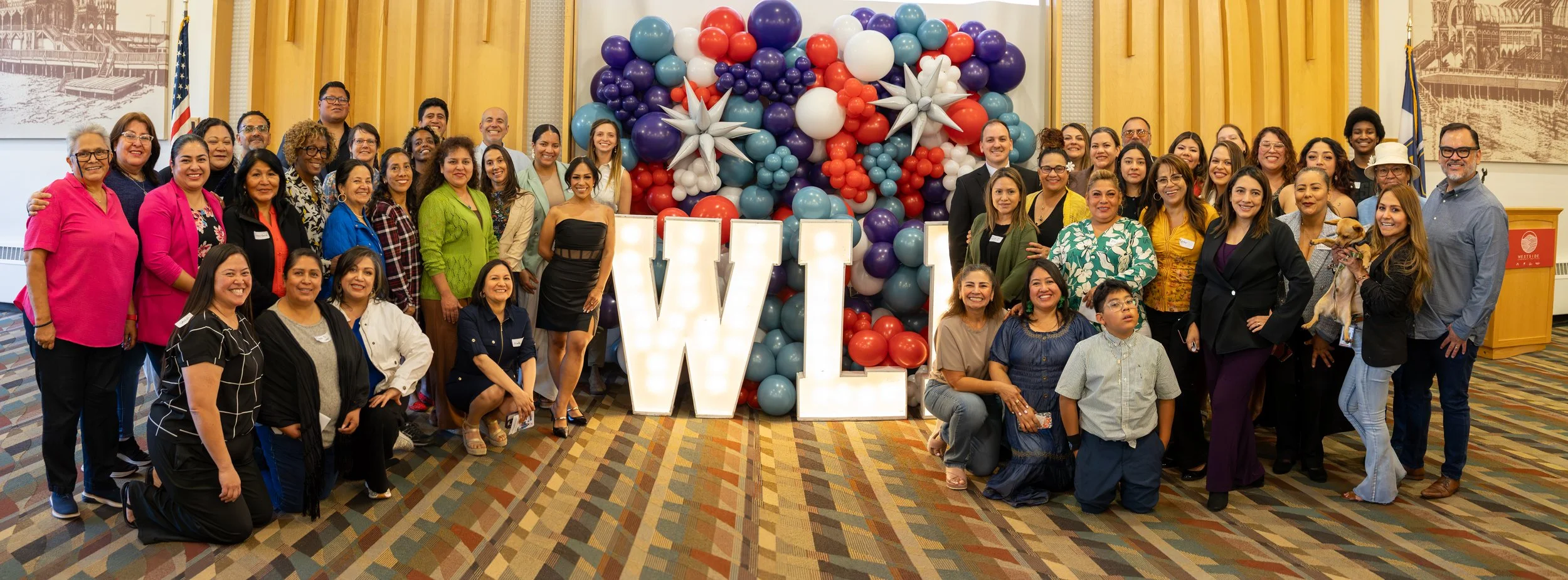 Group of diverse people gathered at an event with a large balloon display and illuminated WEL sign in front of a wall of colorful balloons.