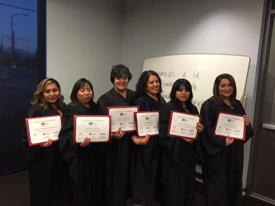 Six women in black graduation robes holding certificates, standing indoors near a whiteboard with writing, in front of a glass window showing an outdoor scene.