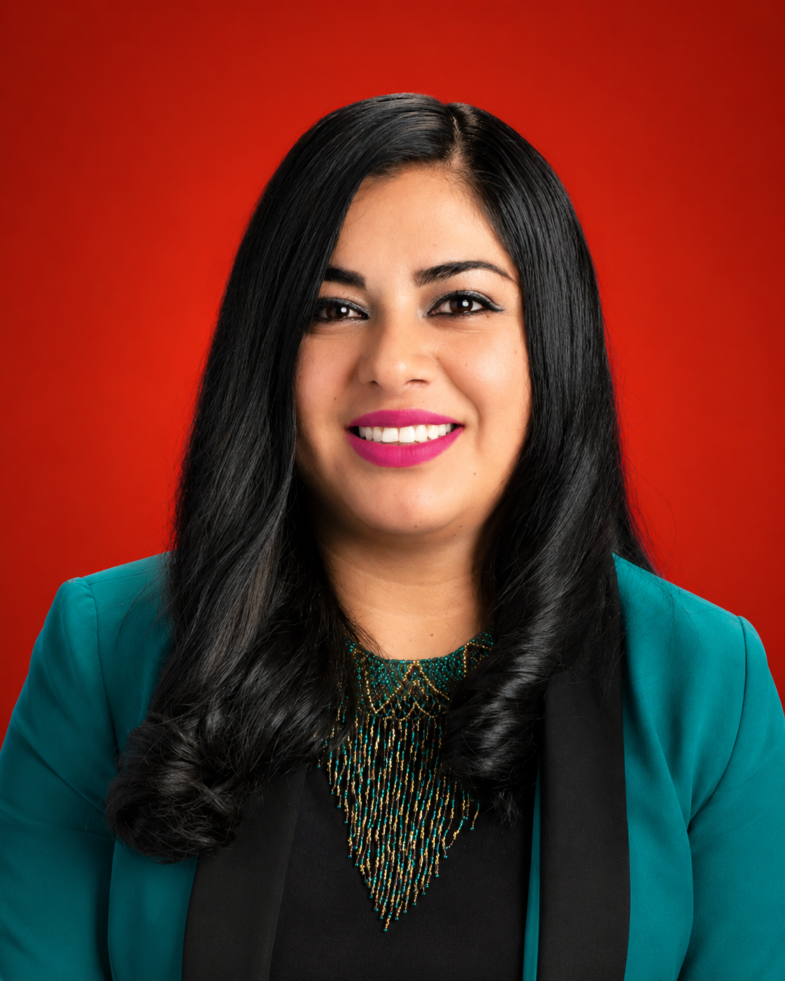 Portrait of a smiling woman with long black hair, wearing a teal blazer and a statement necklace, against a red background.