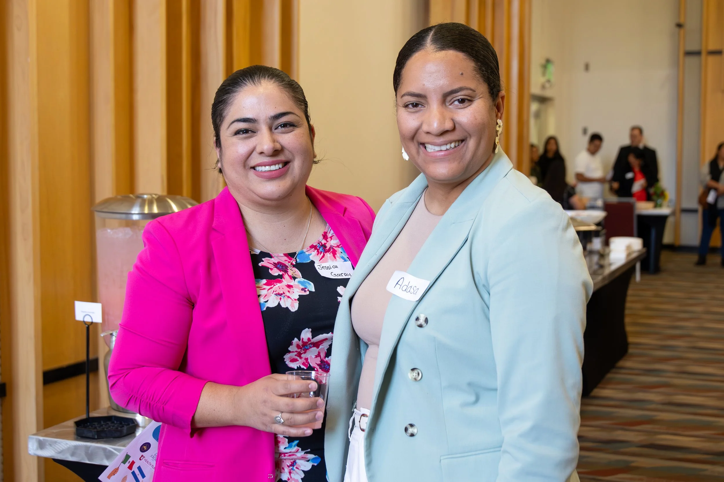 Two women standing close together and smiling at the camera, one in a bright pink blazer and the other in a light green blazer, at a professional or social event.
