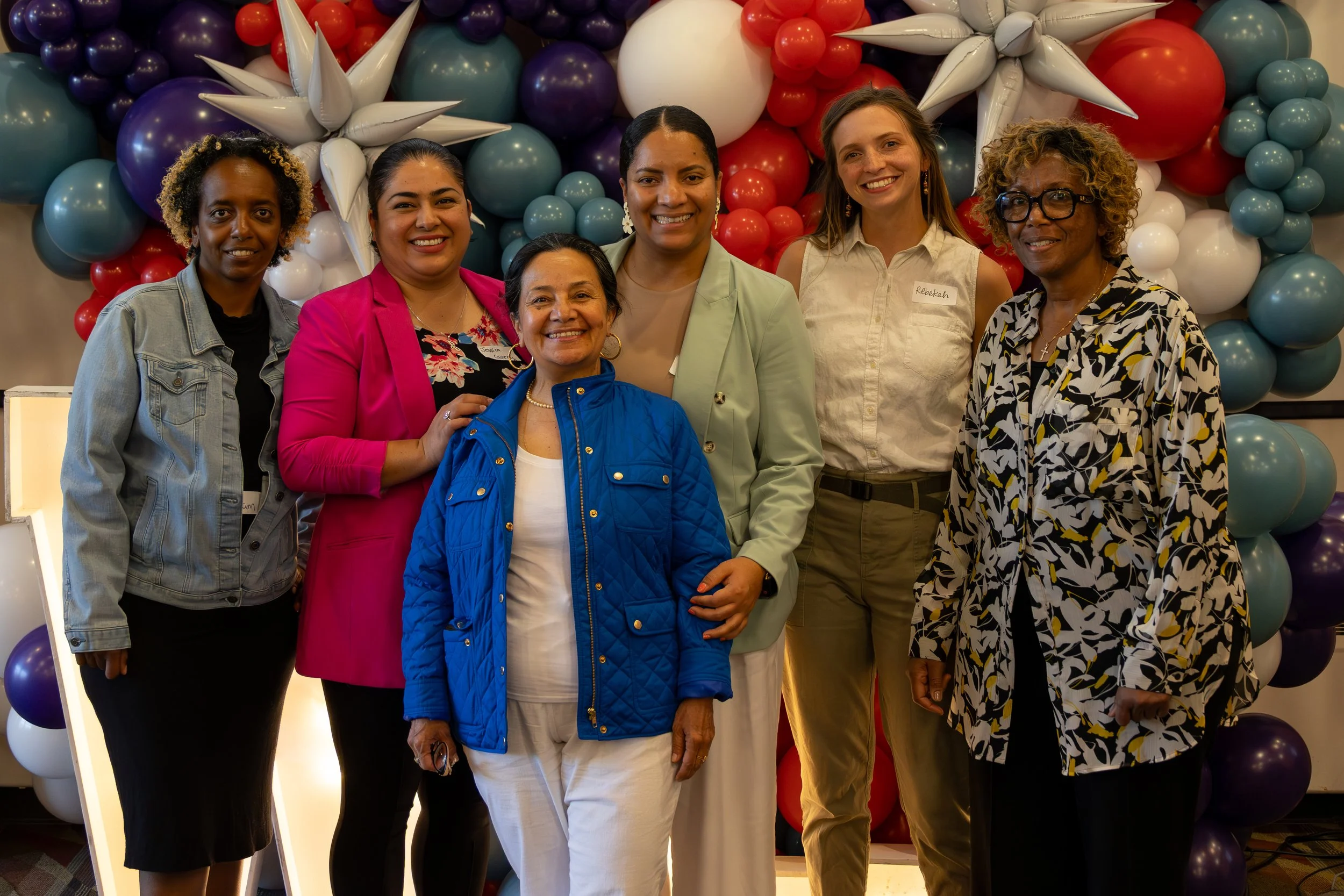 Group of six women standing in front of colorful balloon backdrop at celebration or event.