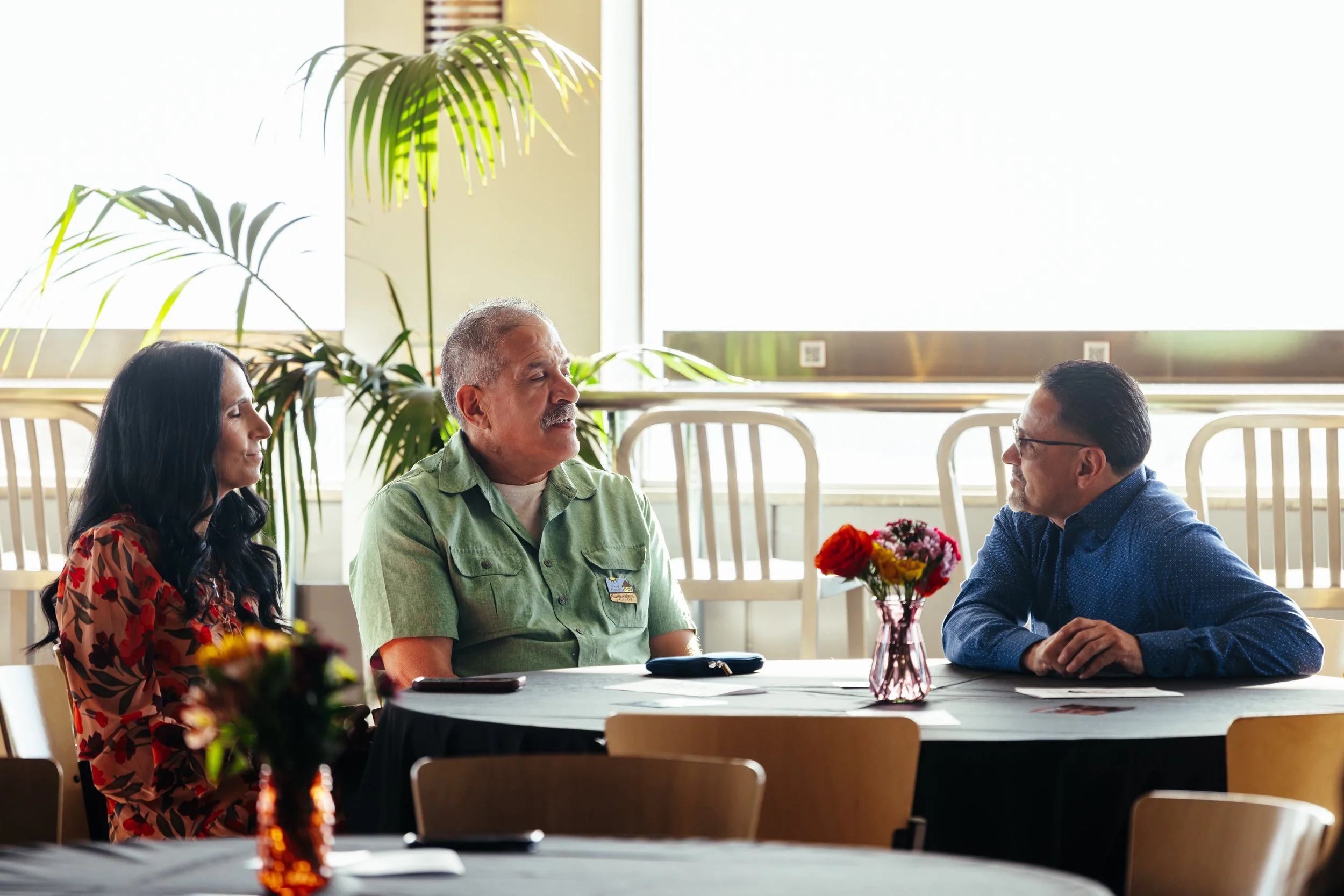 Three people sitting at a round table in a brightly lit room with large windows and green plants in the background, engaged in conversation.