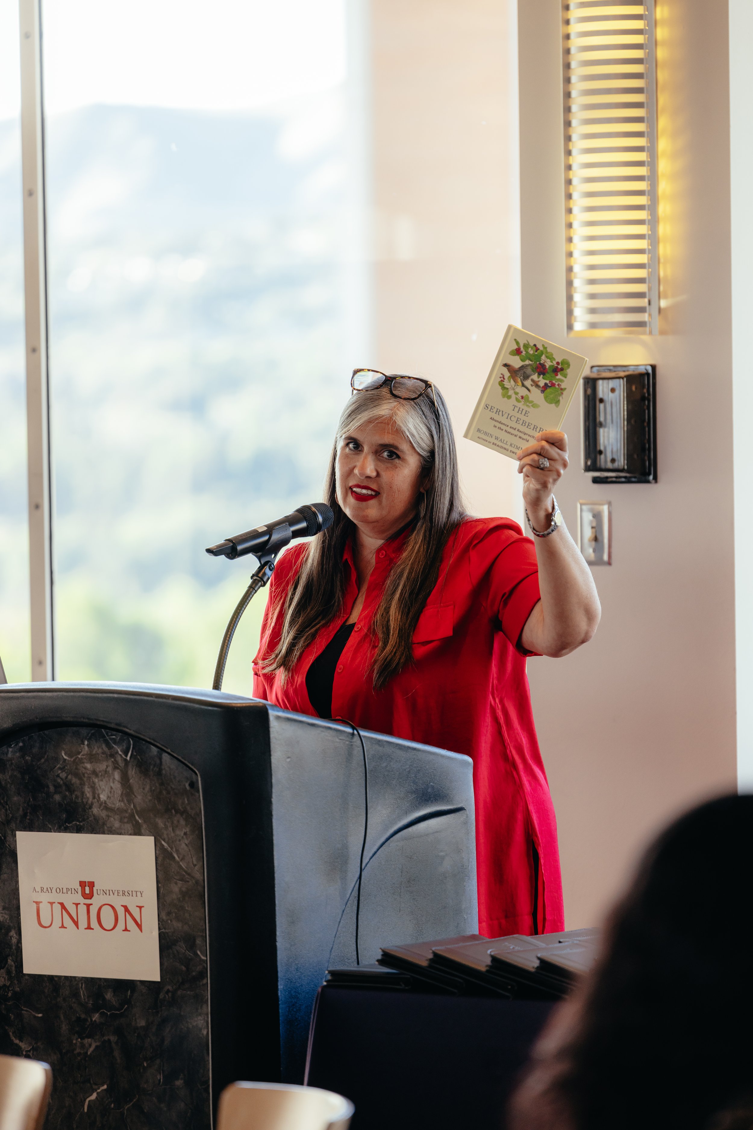 A woman in a red shirt and glasses holding up a book titled 'The Serviceberry' at a podium with a microphone, in a room with large window and yellow lighting.