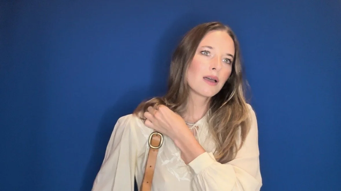 A woman with long brown hair and blue eyes stands against a blue background. She is wearing a light-colored blouse and a tan shoulder bag, looking directly at the camera with a thoughtful expression.