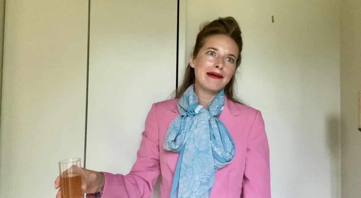 A woman with a surprised expression holding a glass of pinkish beverage, wearing a pink blazer and a large blue scarf, with a plain background.